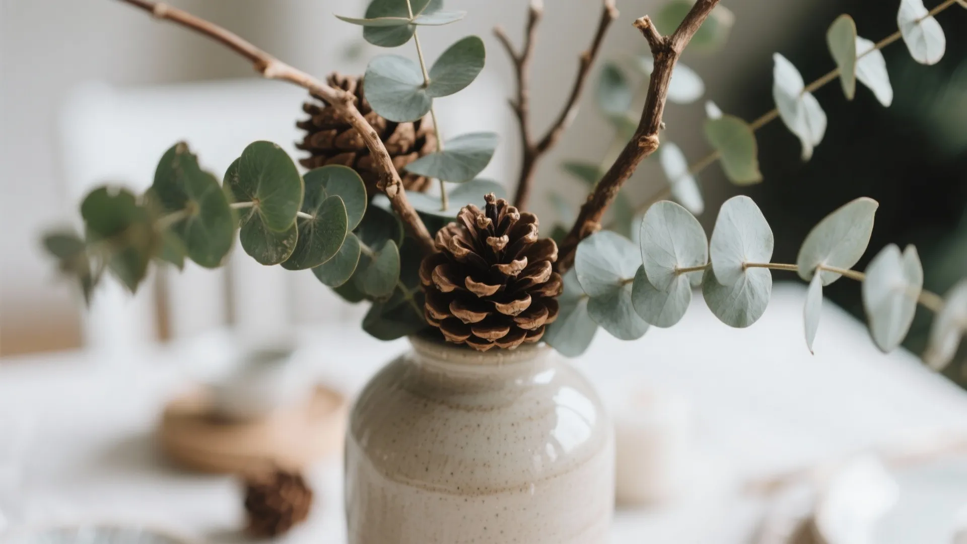 Small ceramic vase holding pine cones and green eucalyptus leaves on a bright white table