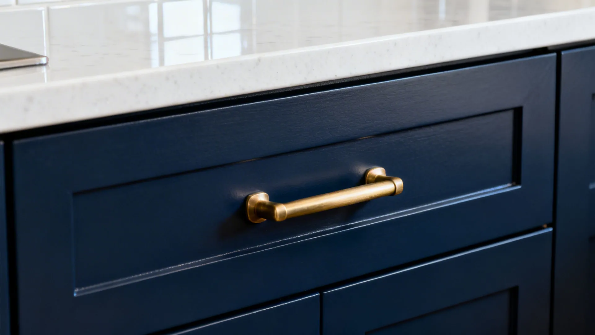Close-up of navy cabinet with brass handle beside white quartz and glossy tile.