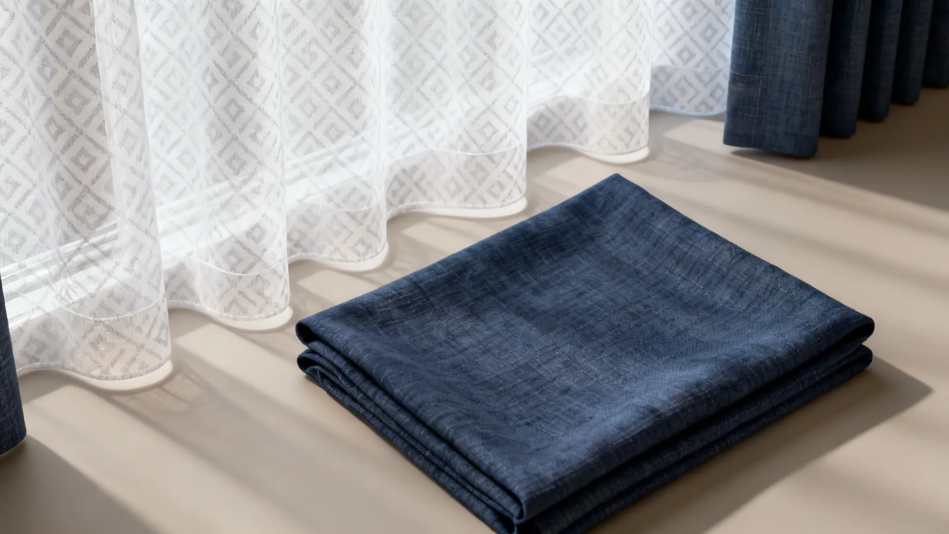 Folded navy blue fabric next to white patterned curtains with sunlight on a light surface