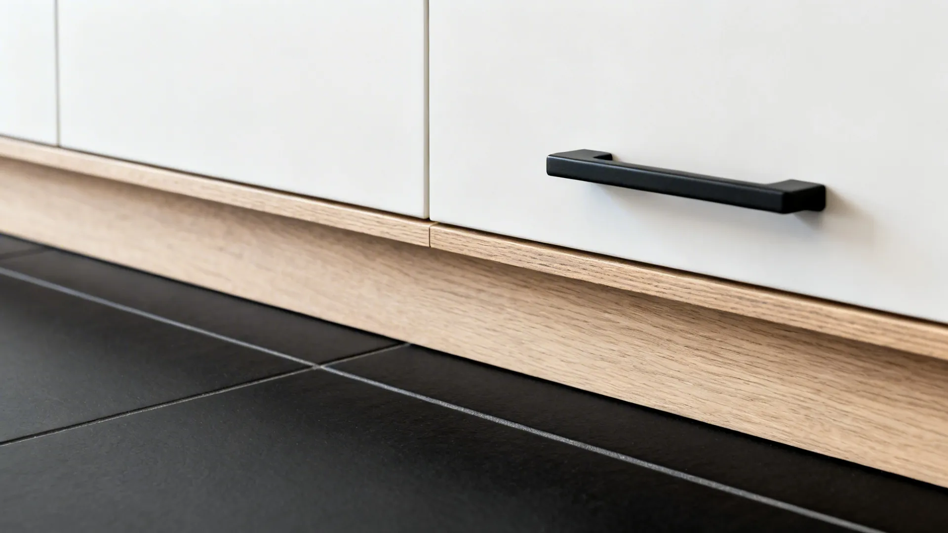 Macro of light oak cabinet toekick meeting a matte black floor with clean grout lines.