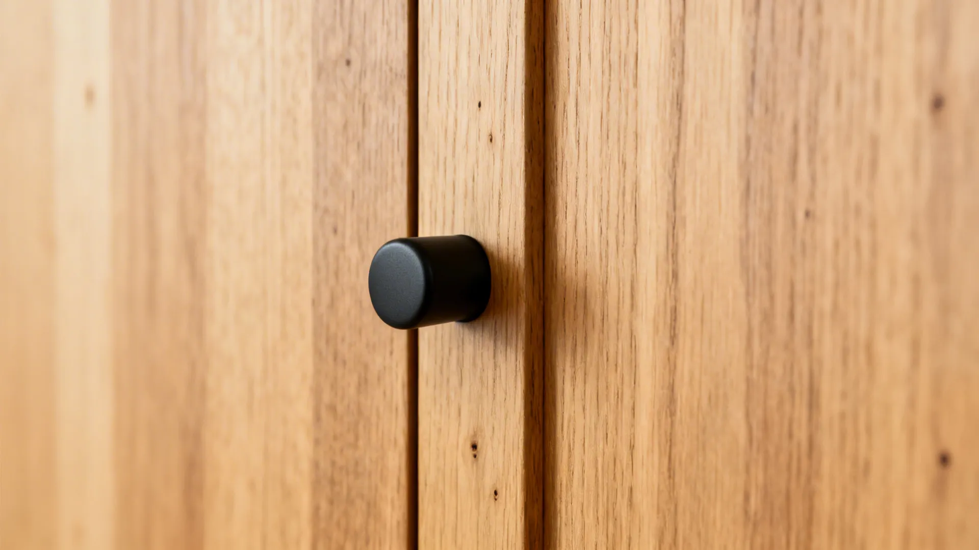 Macro of a vertical-grain light oak cabinet edge with a matte black pull under warm light.