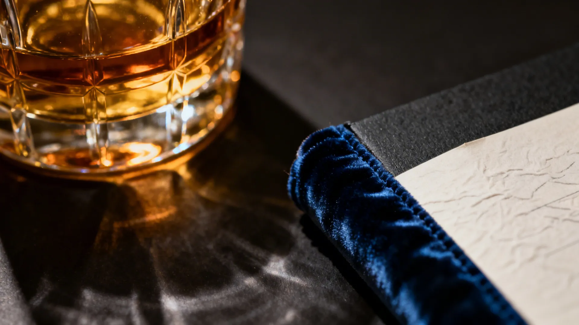 Close-up of amber glass beside ink-blue fabric and a charcoal notebook edge under warm light.