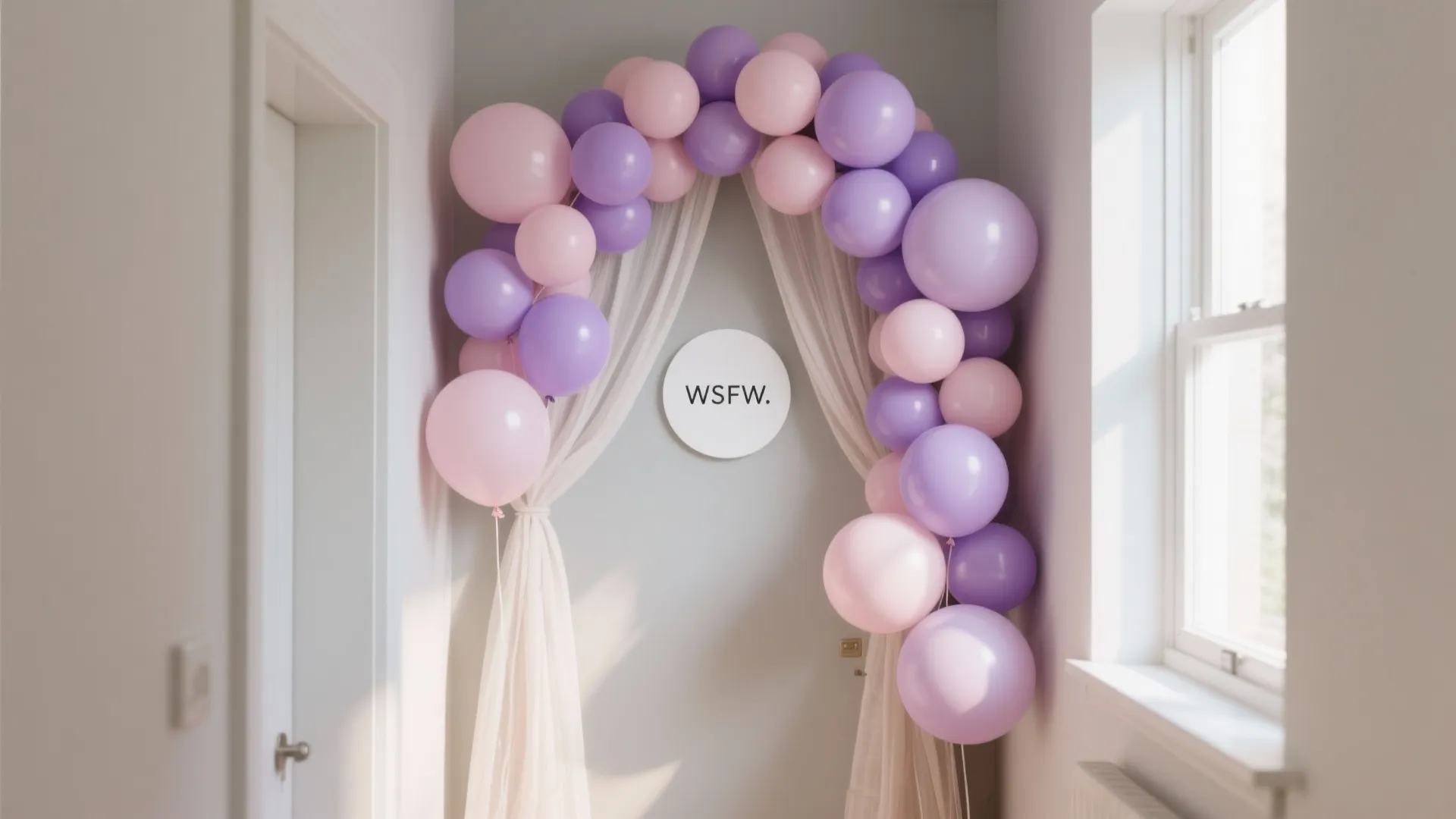 Purple and pink balloon arch over a doorway with white curtains in a bright hallway