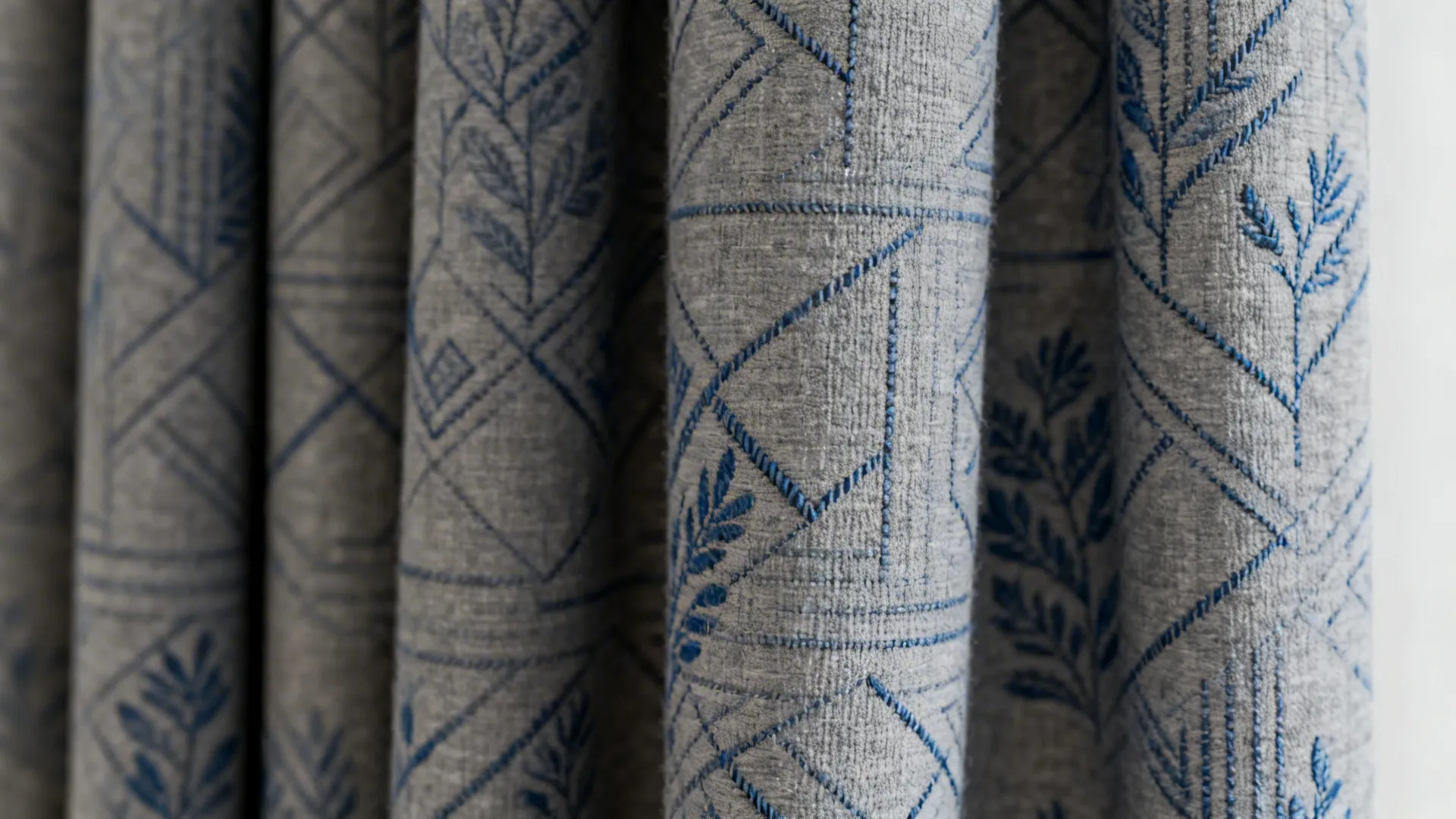 Close-up of grey curtain fabric with subtle blue geometric and botanical motifs showing texture and stitch detail.