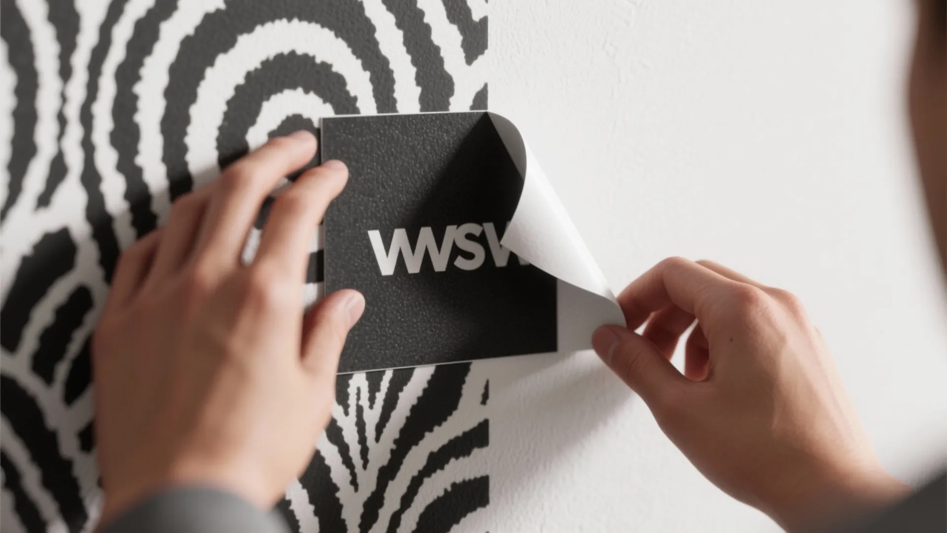 Hands applying a peel-and-stick black-and-white wallpaper swatch to a wall corner
