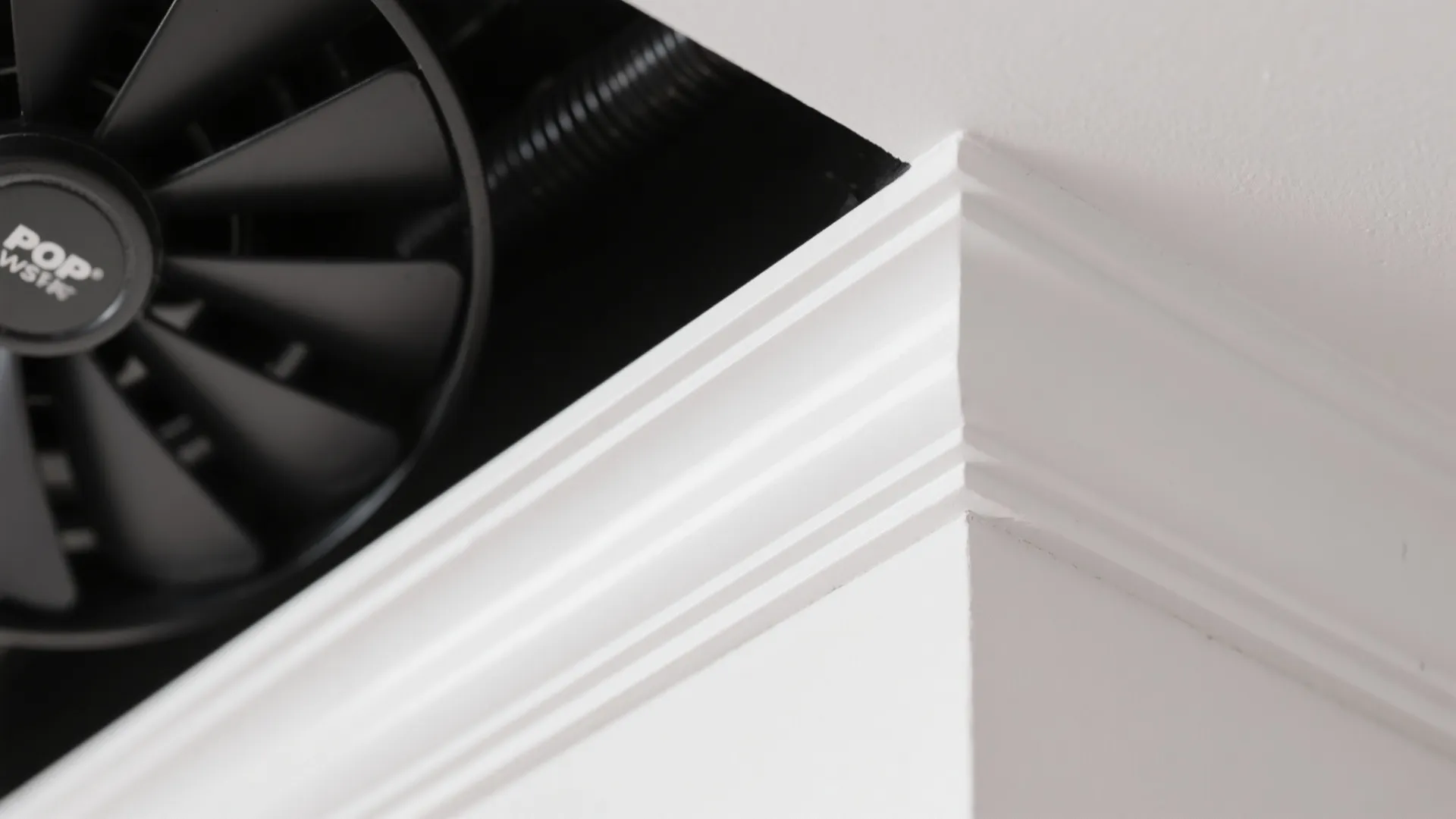 White decorative wall panel corner detail with a black ceiling fan in the background area