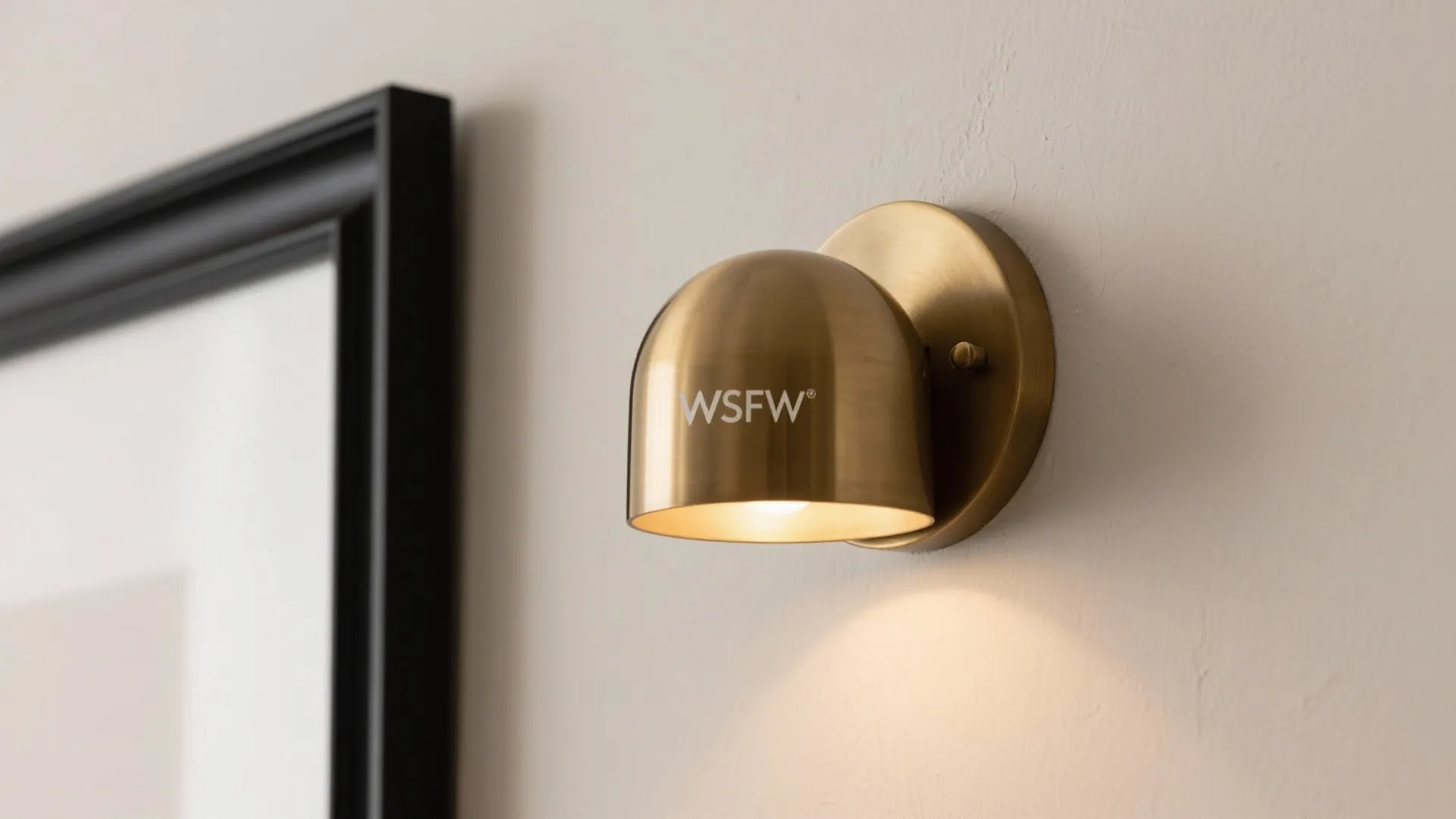 Macro detail of matte black frame edge and brushed brass dome sconce against a putty-painted wall.