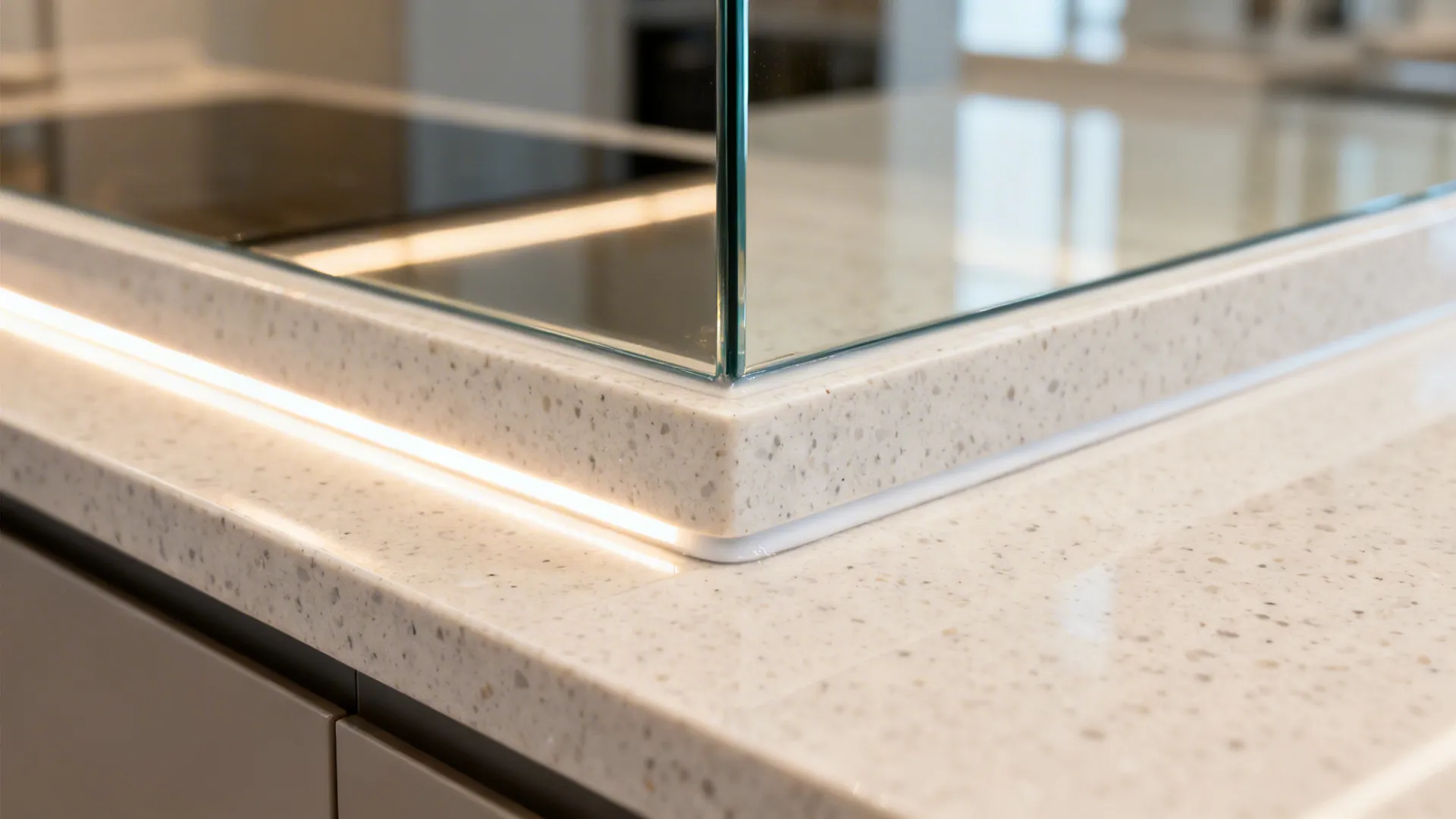 Close-up of light quartz counter meeting reflective glass backsplash under warm LED light.