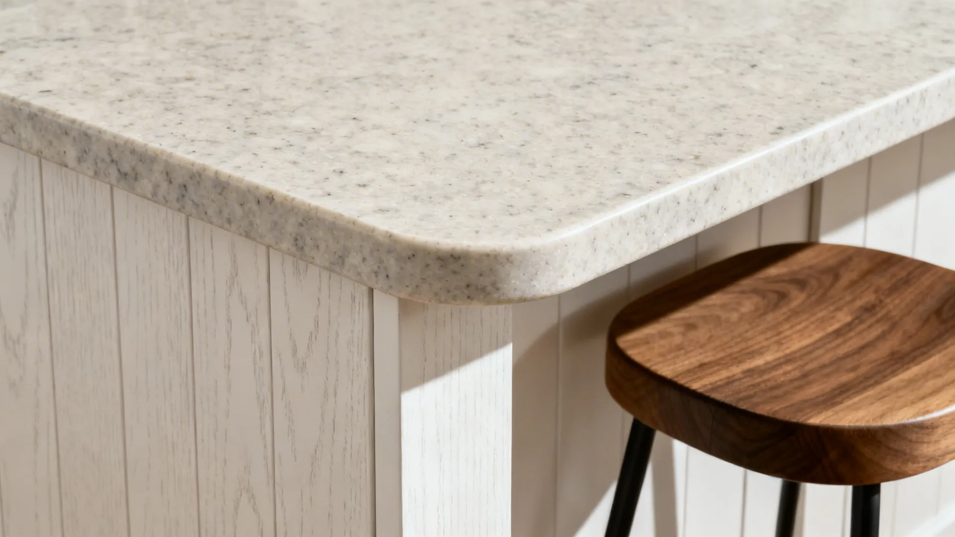 Close up of rounded stone countertop edge on white wooden kitchen island beside wooden stool