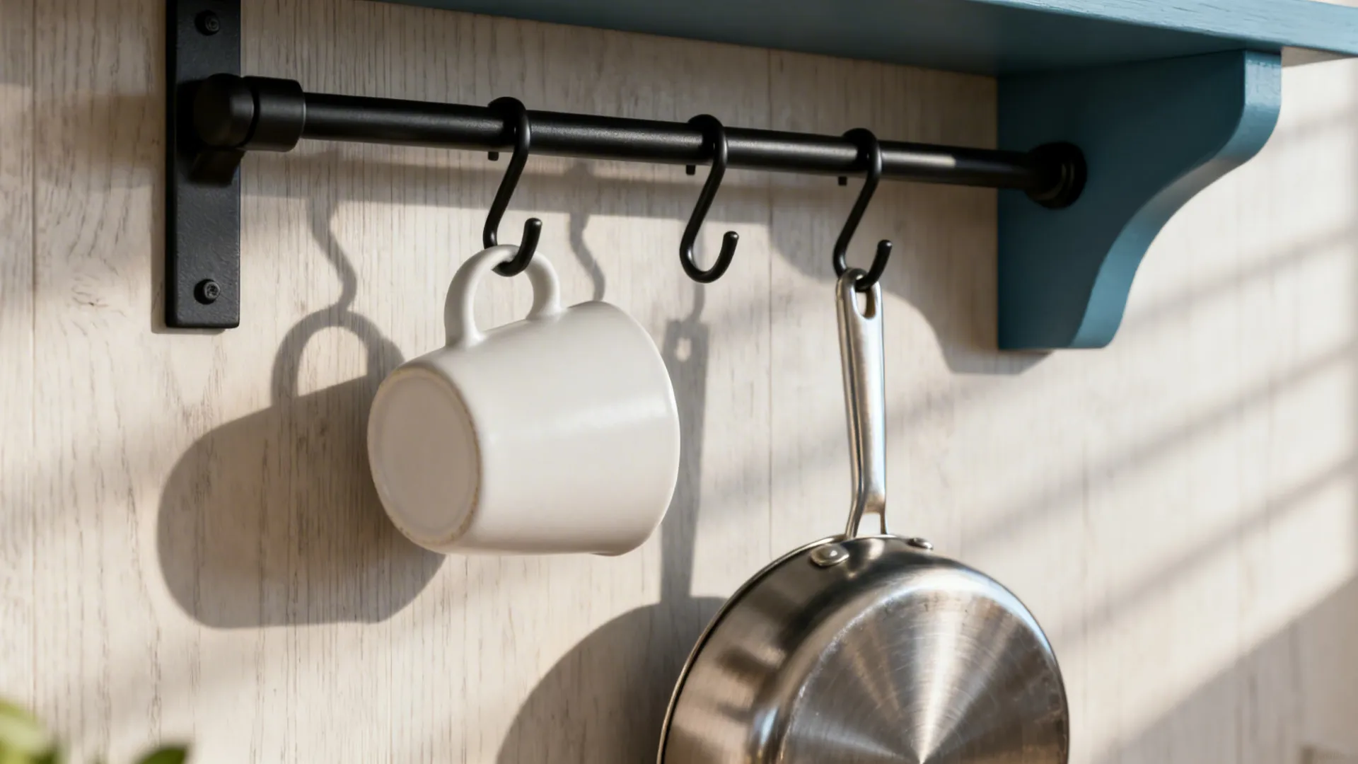 Close-up of black kitchen rail with hooks holding a mug and pan against a neutral wall.