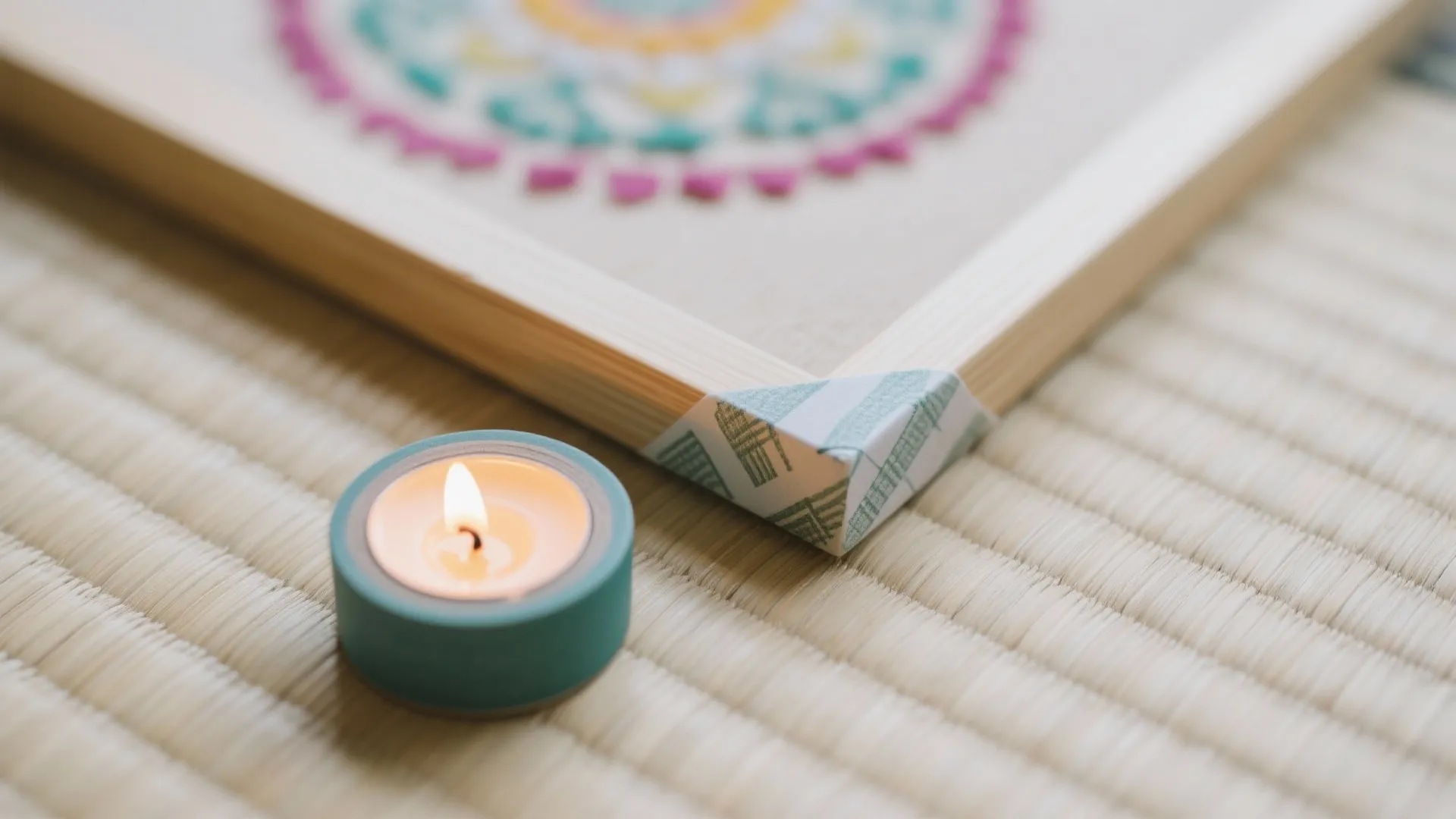 Lit candle in teal holder next to wooden frame with colorful pattern on straw mat