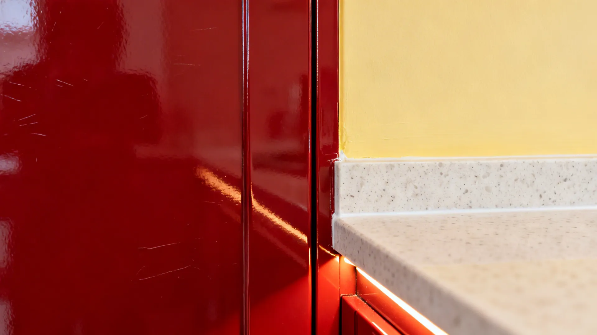 Macro of glossy red cabinet meeting light quartz and soft yellow wall under warm task light.