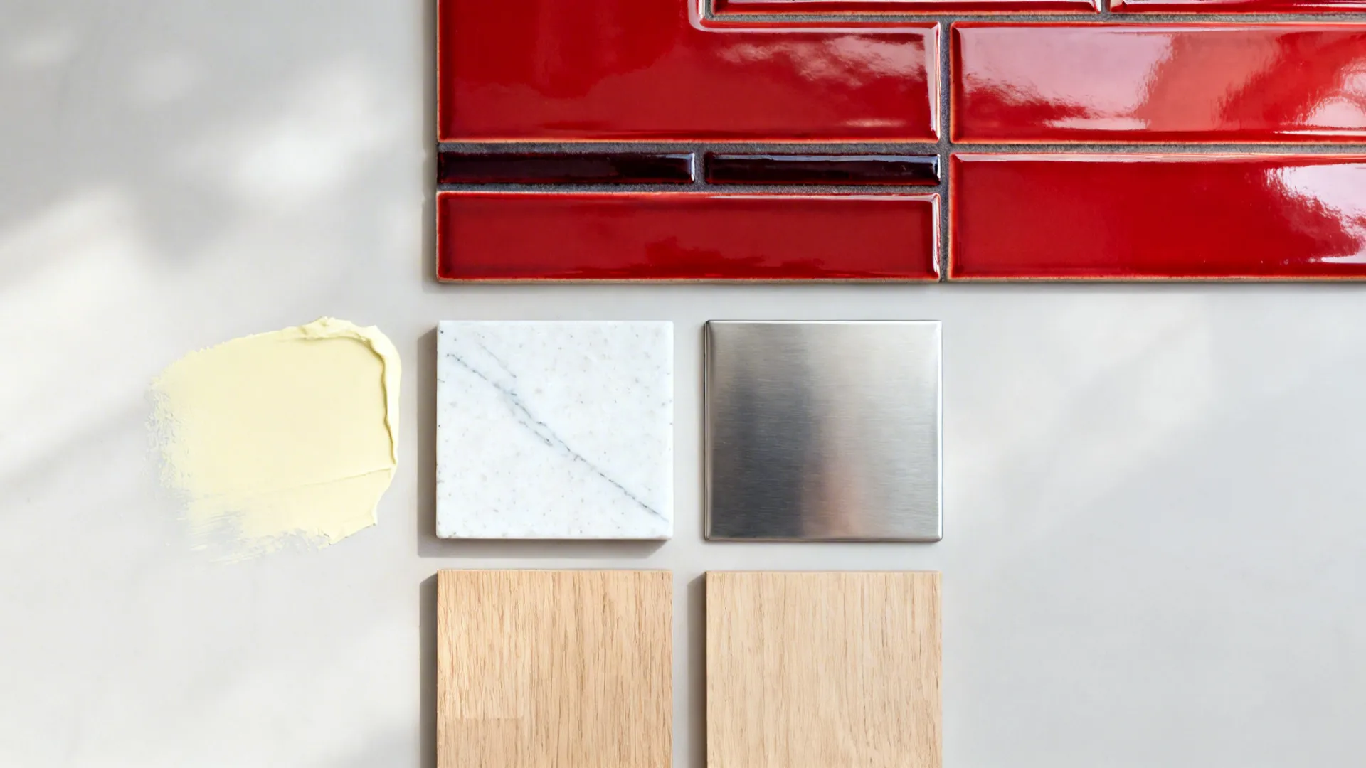Flatlay of glossy red tile, pale yellow paint, white quartz, stainless, and light oak samples.