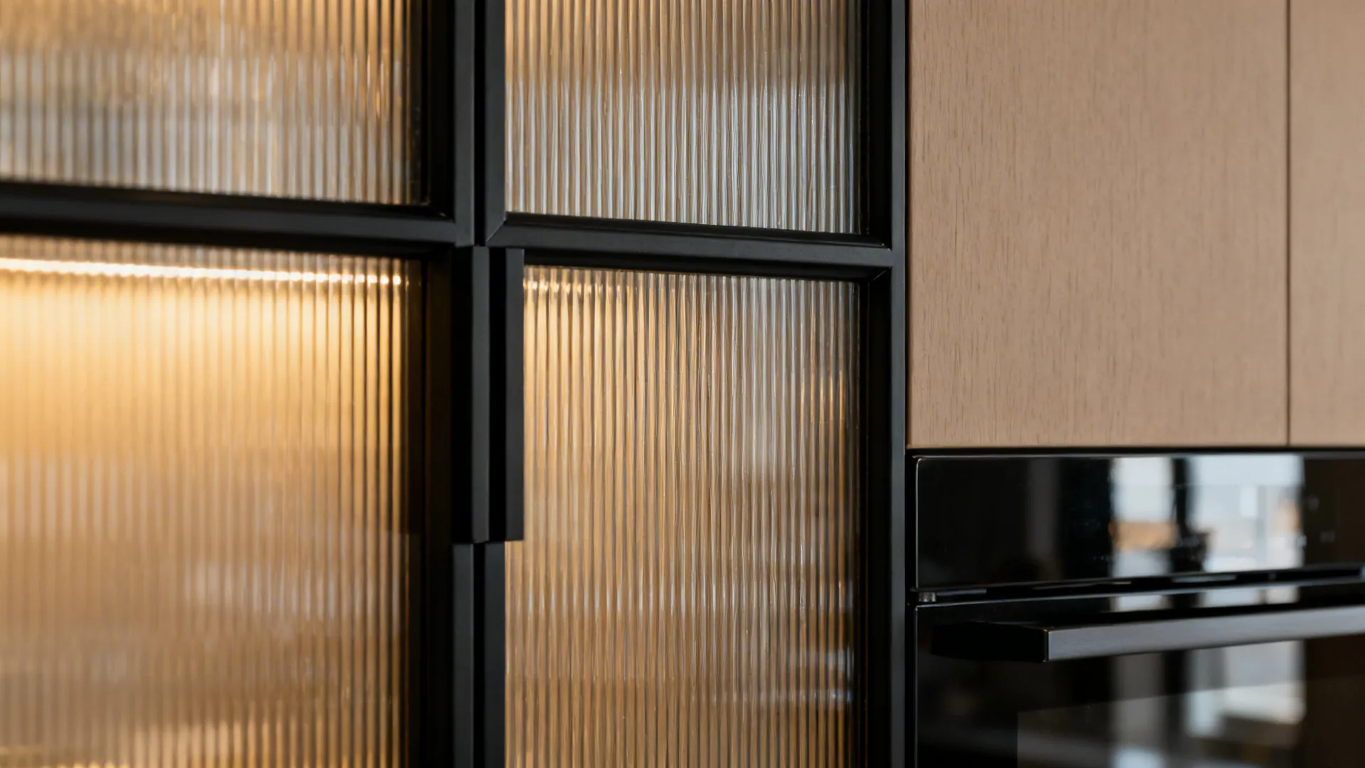 Close-up of black-framed reeded glass next to taupe cabinetry and a satin black appliance edge.
