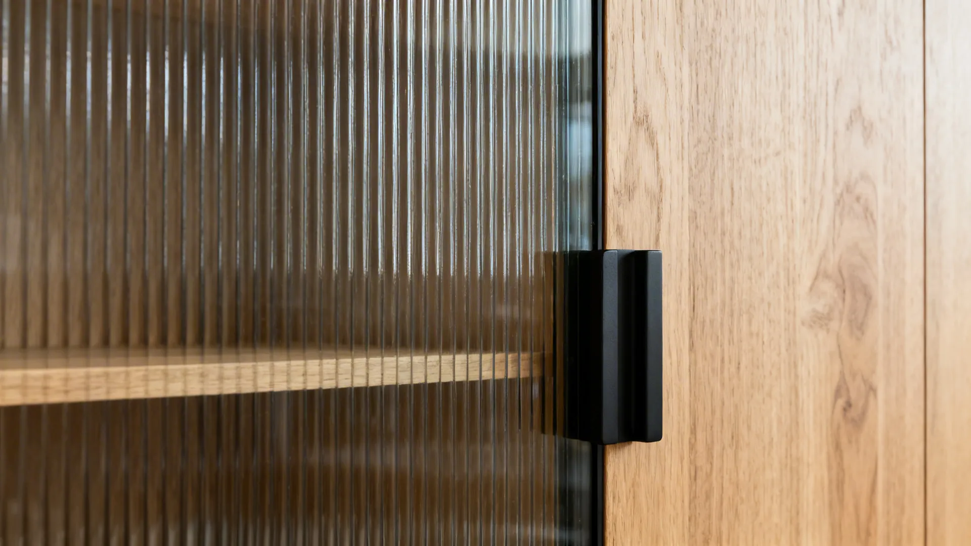 Macro detail of ribbed glass cabinet door, light oak shelving, and a matte black handle in soft daylight.