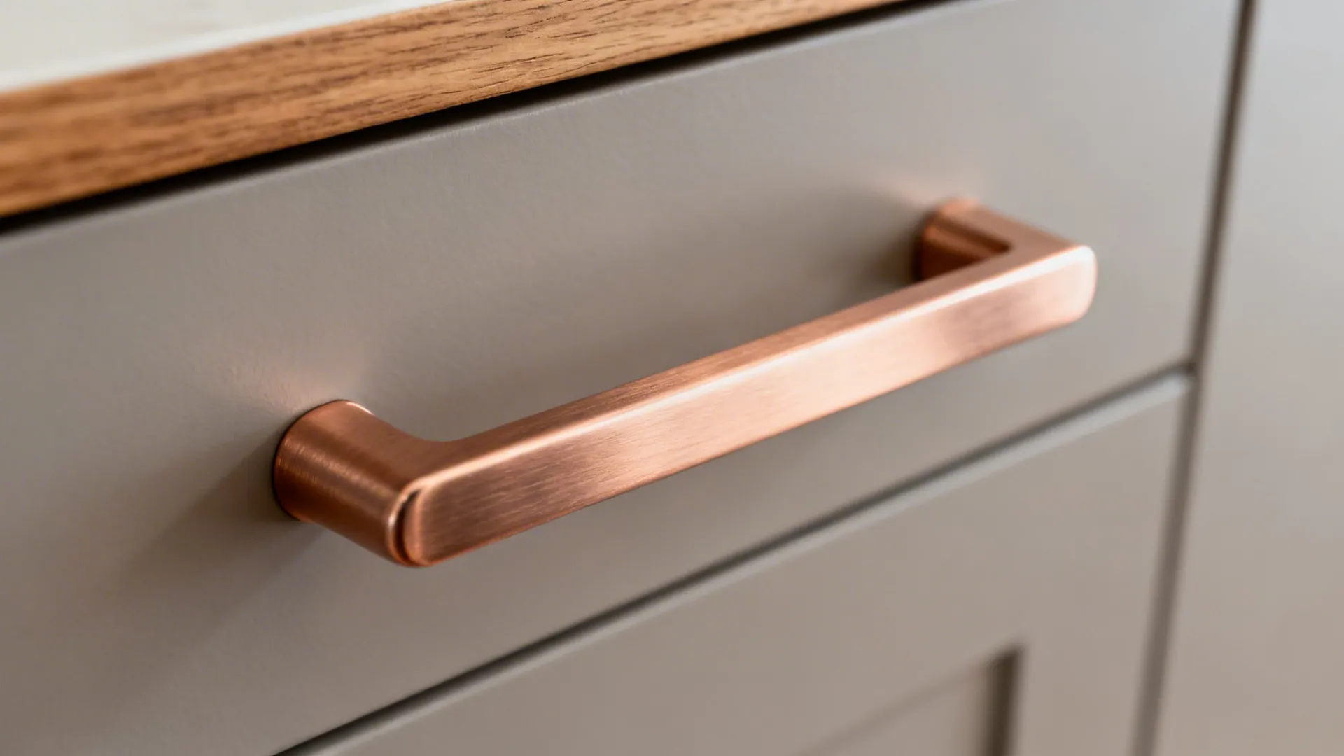 Macro of brushed rose gold cabinet pull on matte drawer with oak detail in soft daylight.