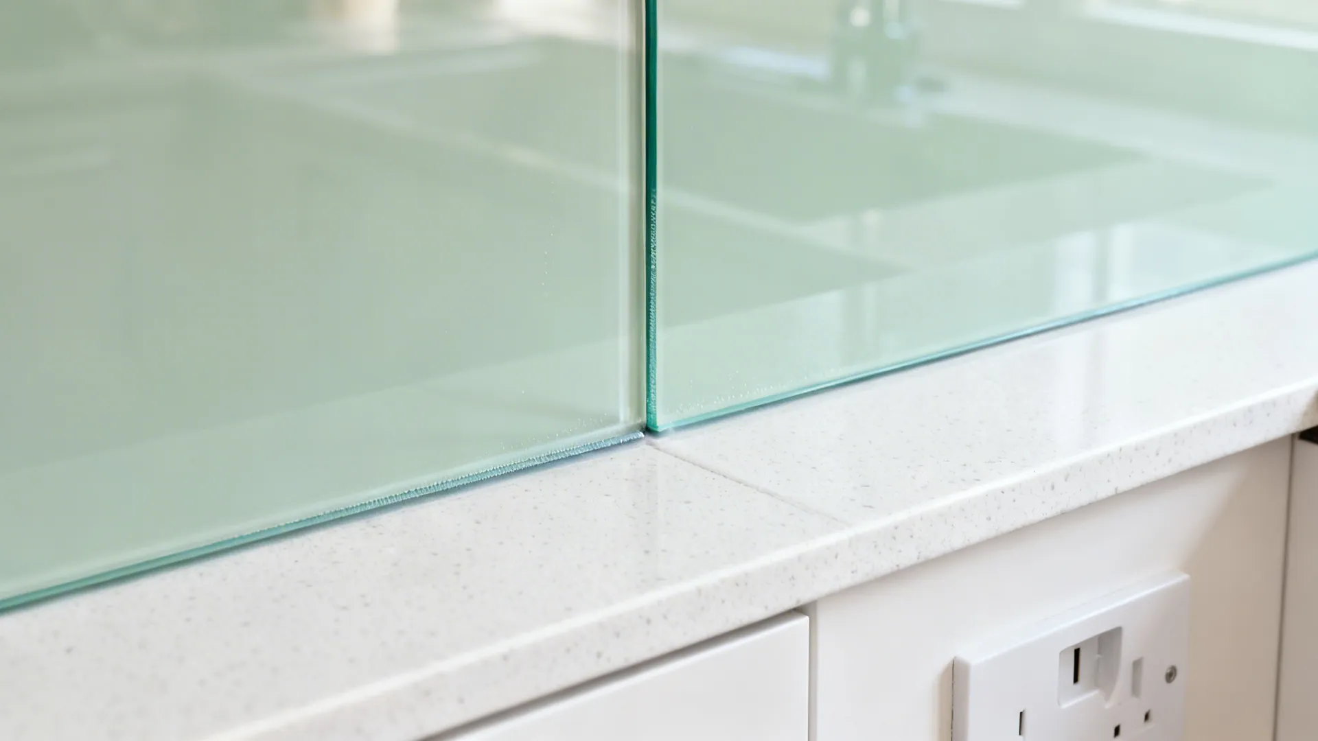 Macro of a pale sage back-painted glass splashback meeting a white quartz worktop in soft daylight.