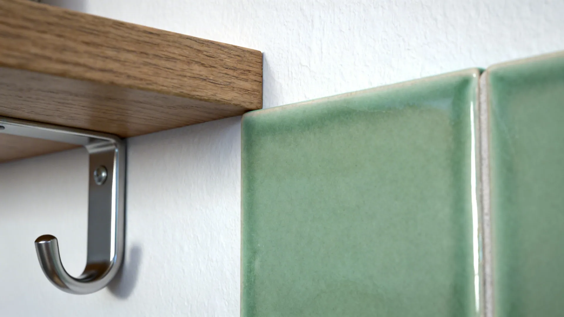 Macro of sage ceramic tile beside soft white paint and oak shelf with a matte nickel hook.
