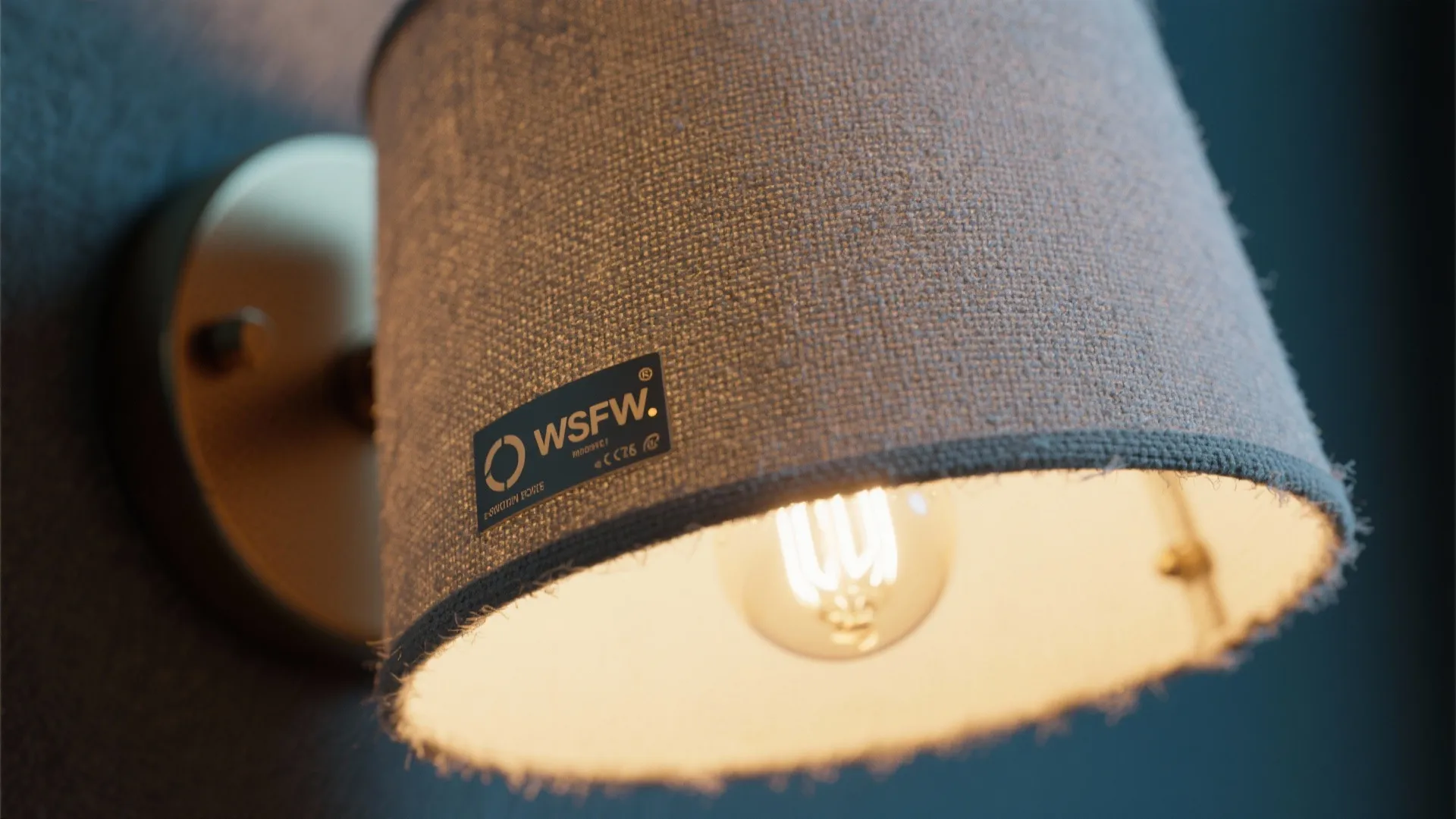 Macro detail of an angled fabric sconce showing fabric weave, warm bulb glow, and moisture-rated marking.