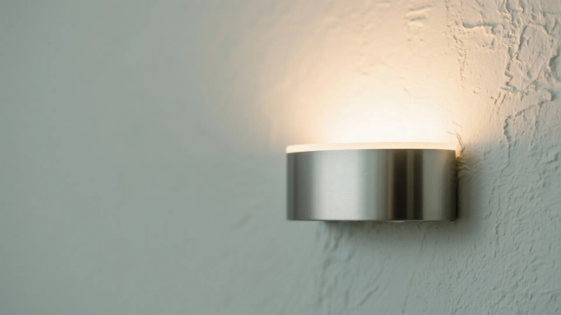 Close up of a round silver wall light fixture glowing on a white textured wall surface