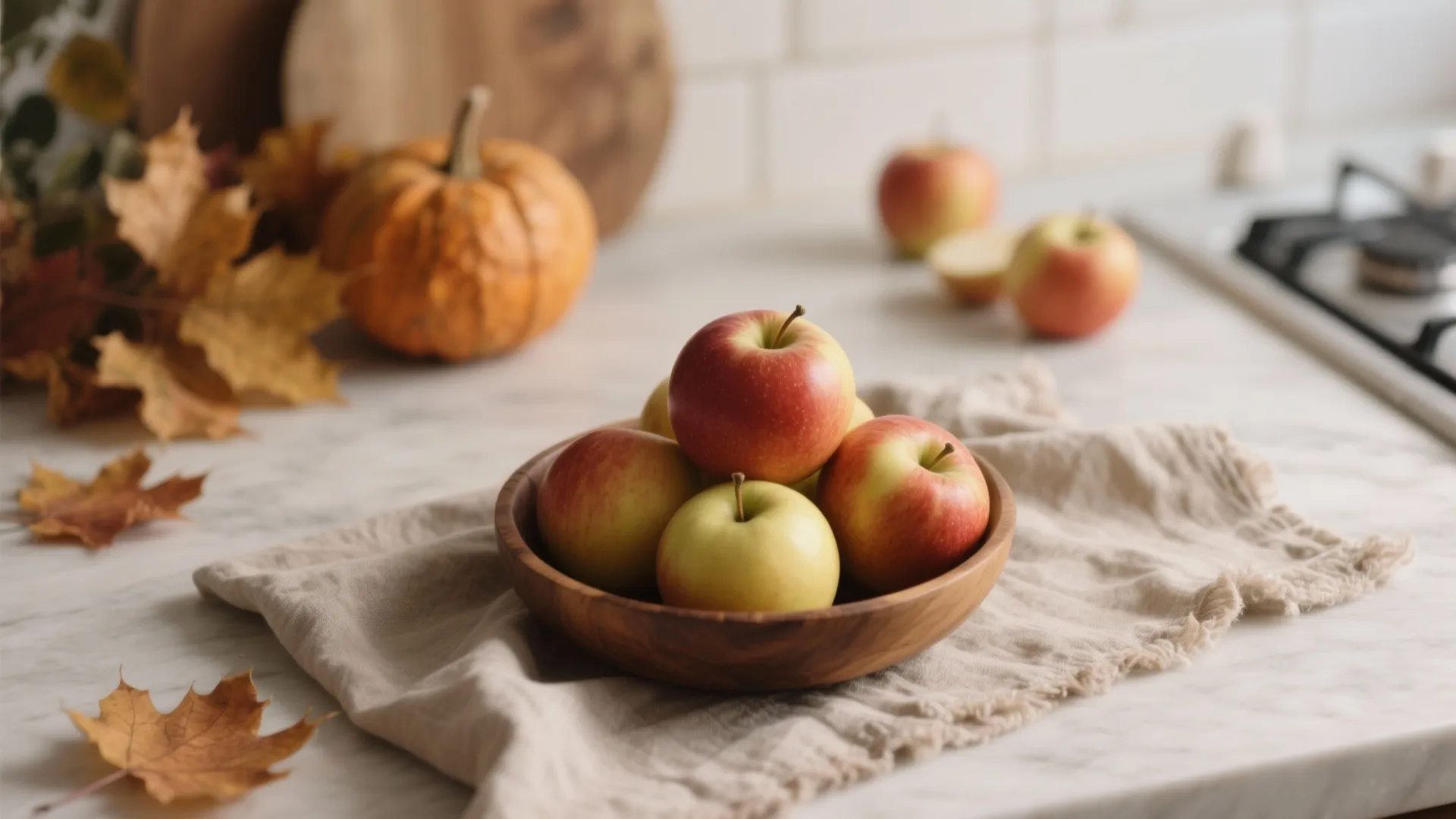 Wooden bowl filled with red apples next to a pumpkin and dry autumn maple leaves
