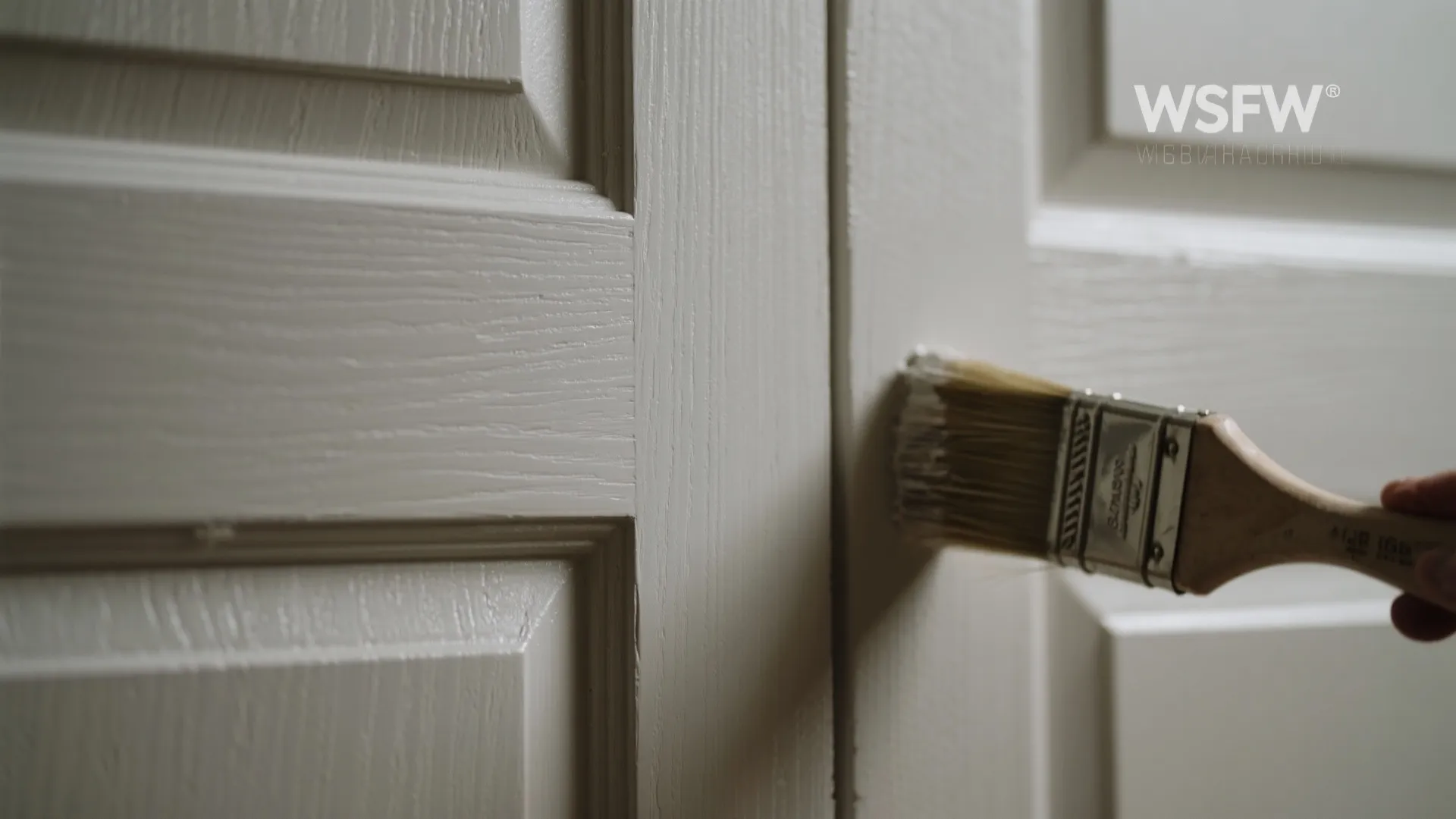 Close up view of a paint brush applying light grey paint to a wooden wall panel