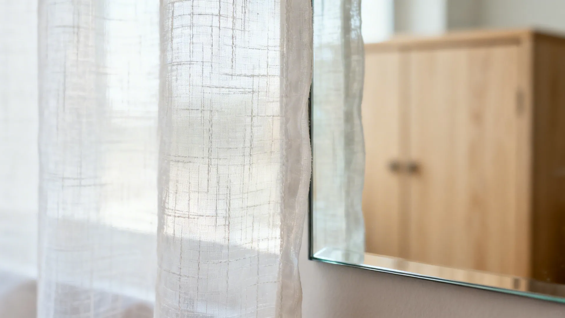 Close-up of airy sheer fabric beside a clean mirror edge reflecting a light wood cabinet.