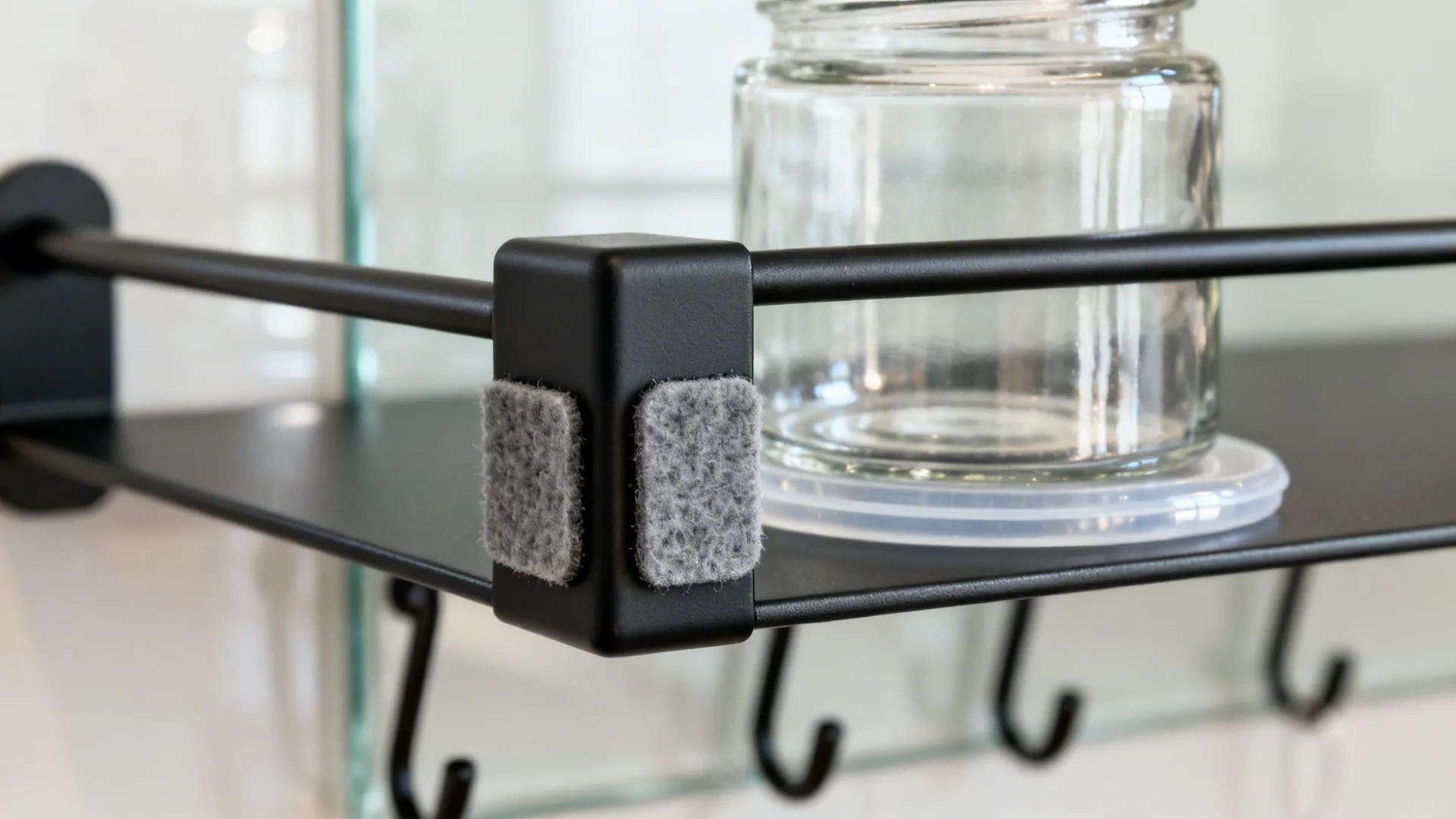 Black metal shelf corner with gray pads holding a glass jar above several small hooks