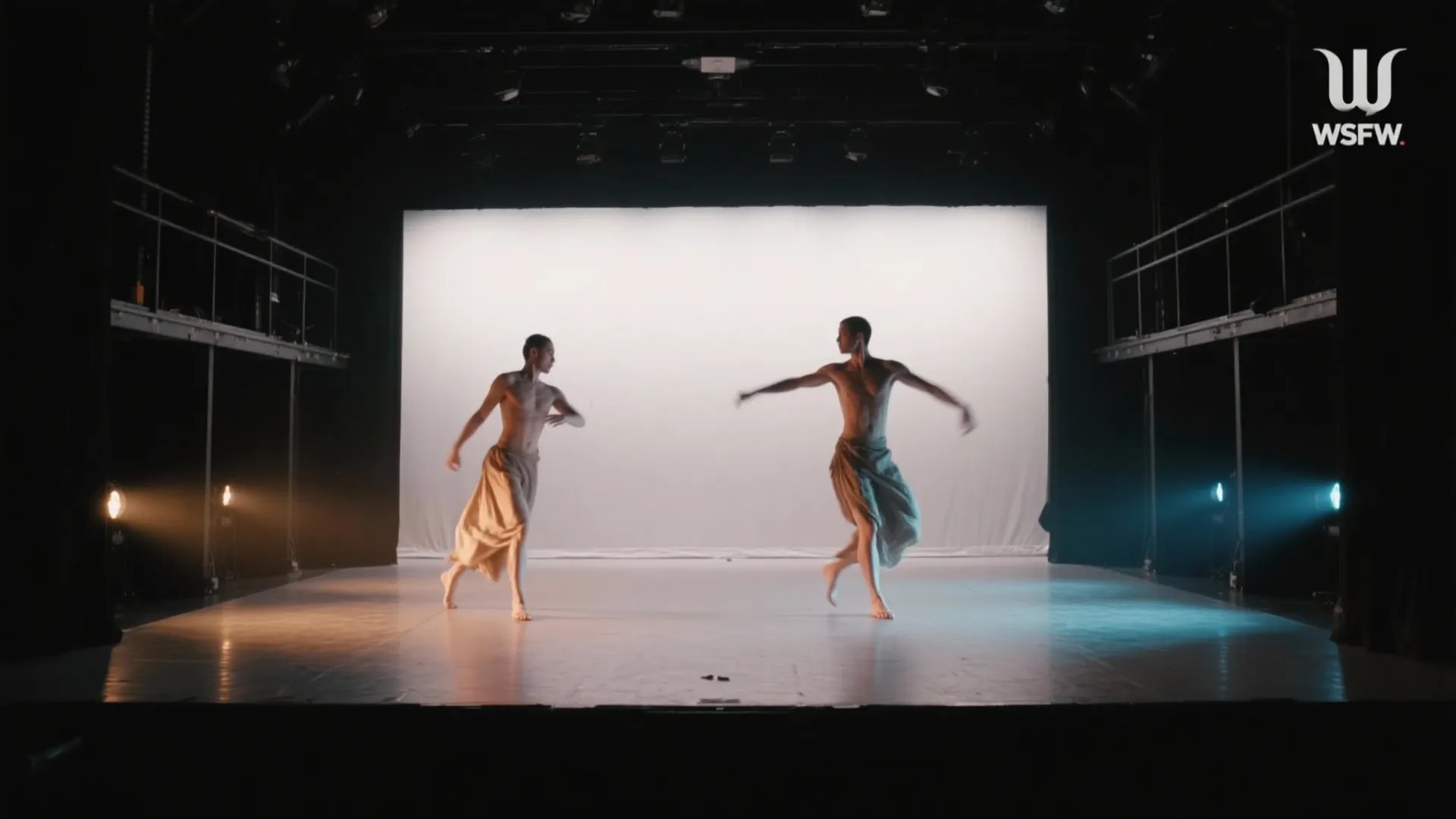 Two male dancers performing on a brightly lit stage with white background and side light fixtures