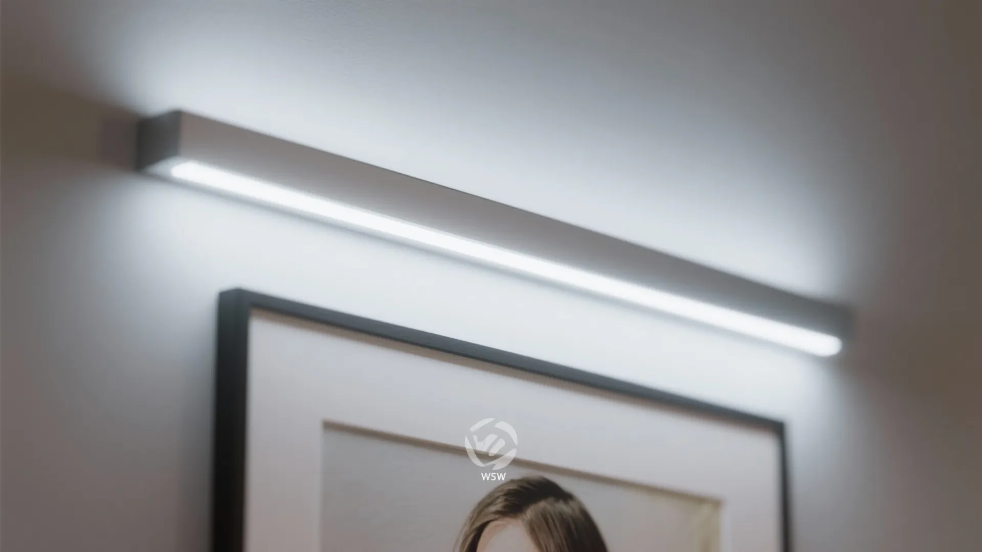 Modern long white light fixture mounted on a wall above a black framed picture frame