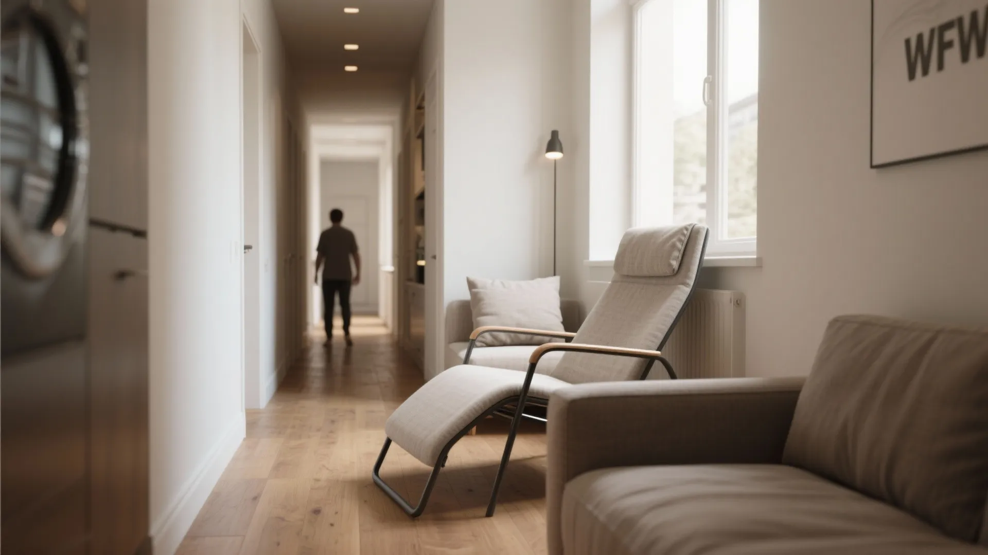 Narrow hallway with wooden floor featuring a grey sofa and person walking in the background