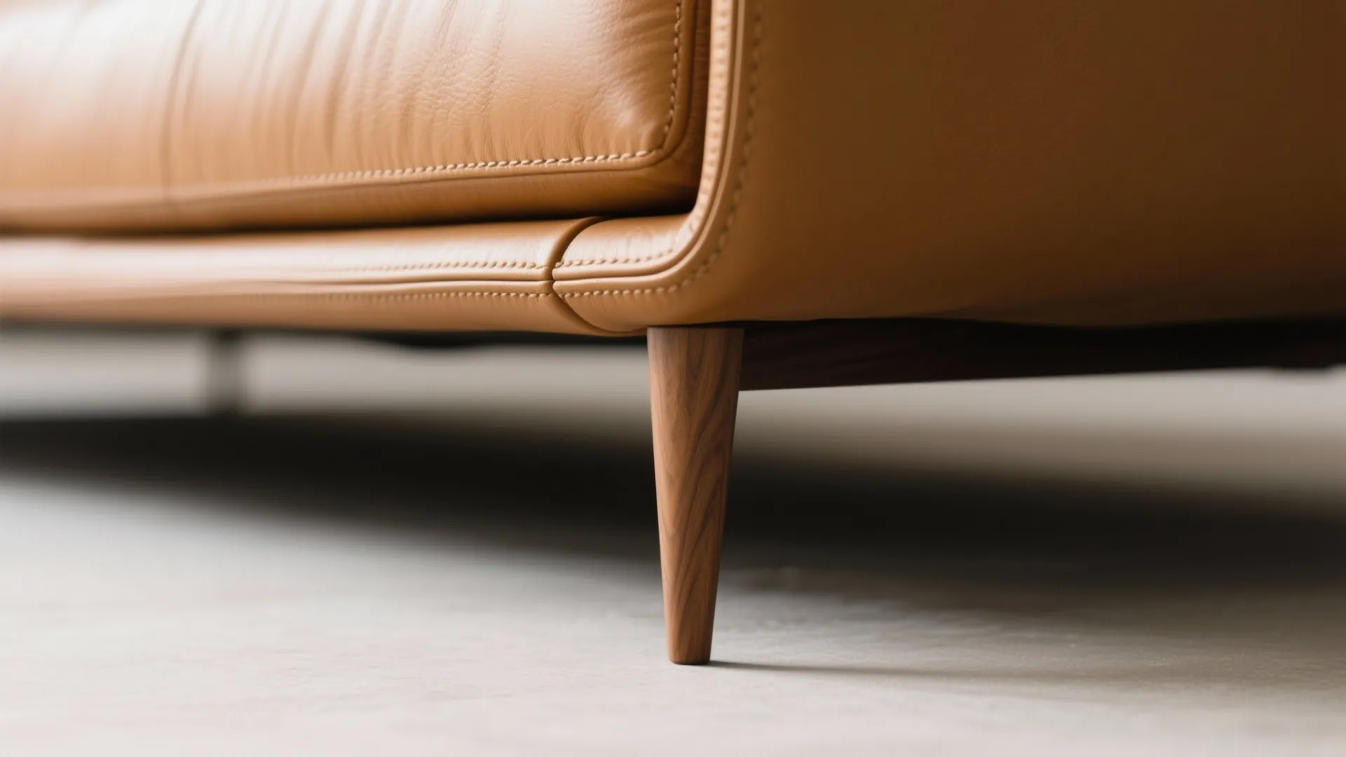 Close up view of light brown leather sofa with wooden legs and detailed stitching on side