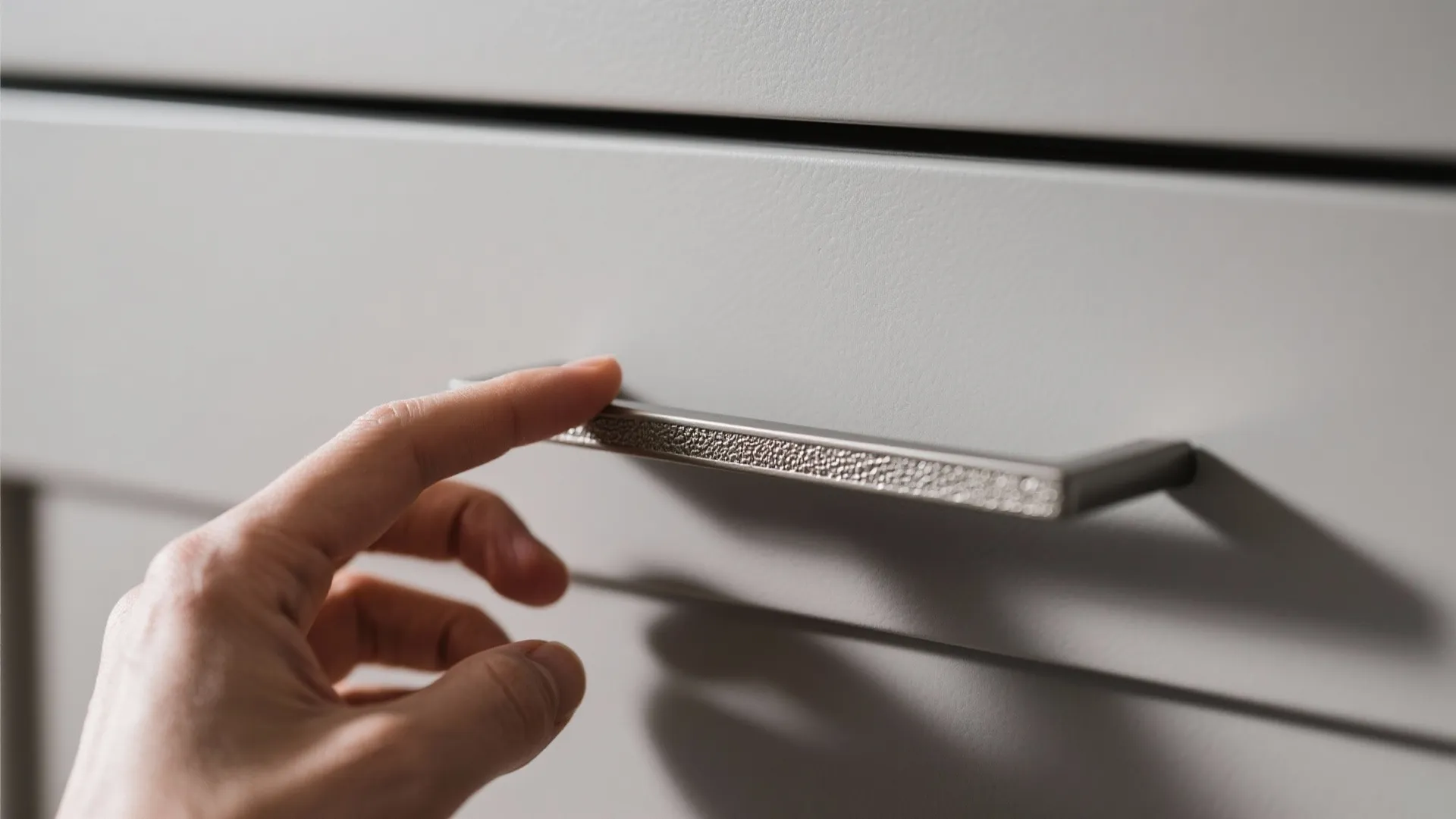Person's hand touching a textured silver metal handle on a light grey kitchen cabinet drawer