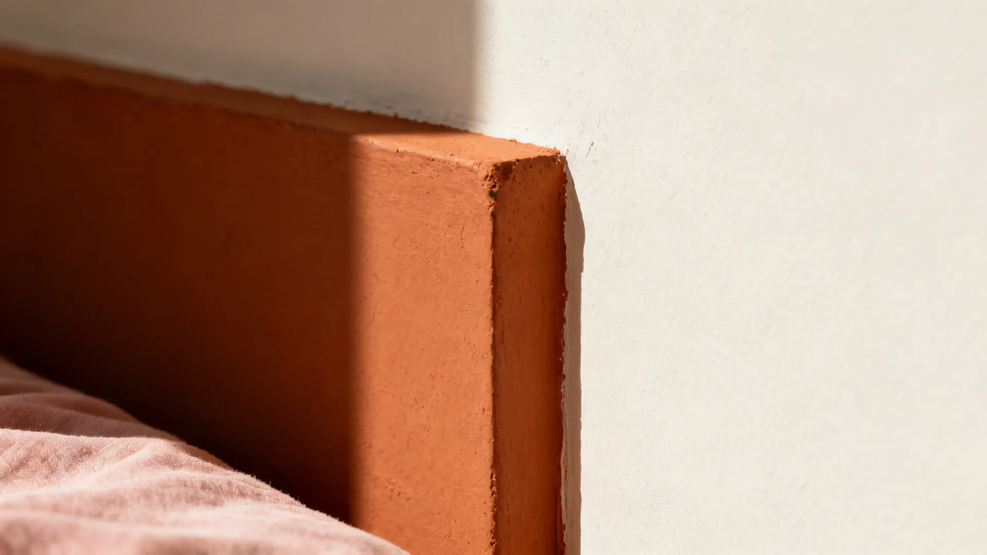 Macro of terracotta paint band meeting off-white wall with warm light and blush fabric detail.