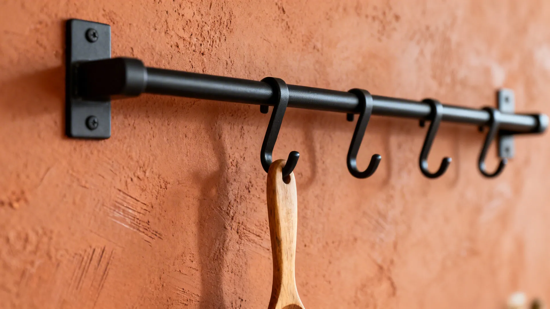 Macro of terracotta wall with a matte black kitchen rail and wooden utensil in warm light.