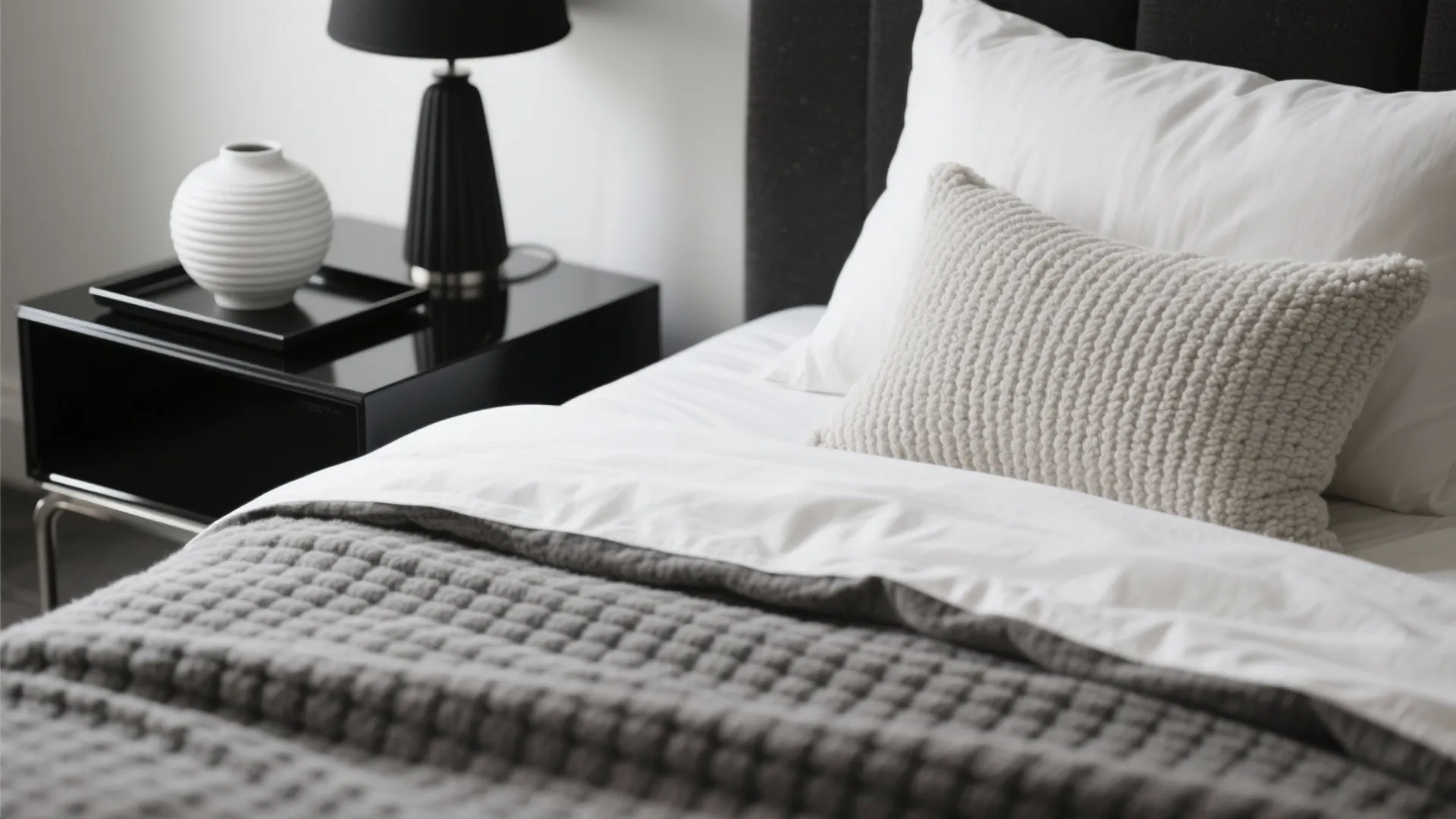 Close up of bed with white pillows textured grey blanket black nightstand and white vase