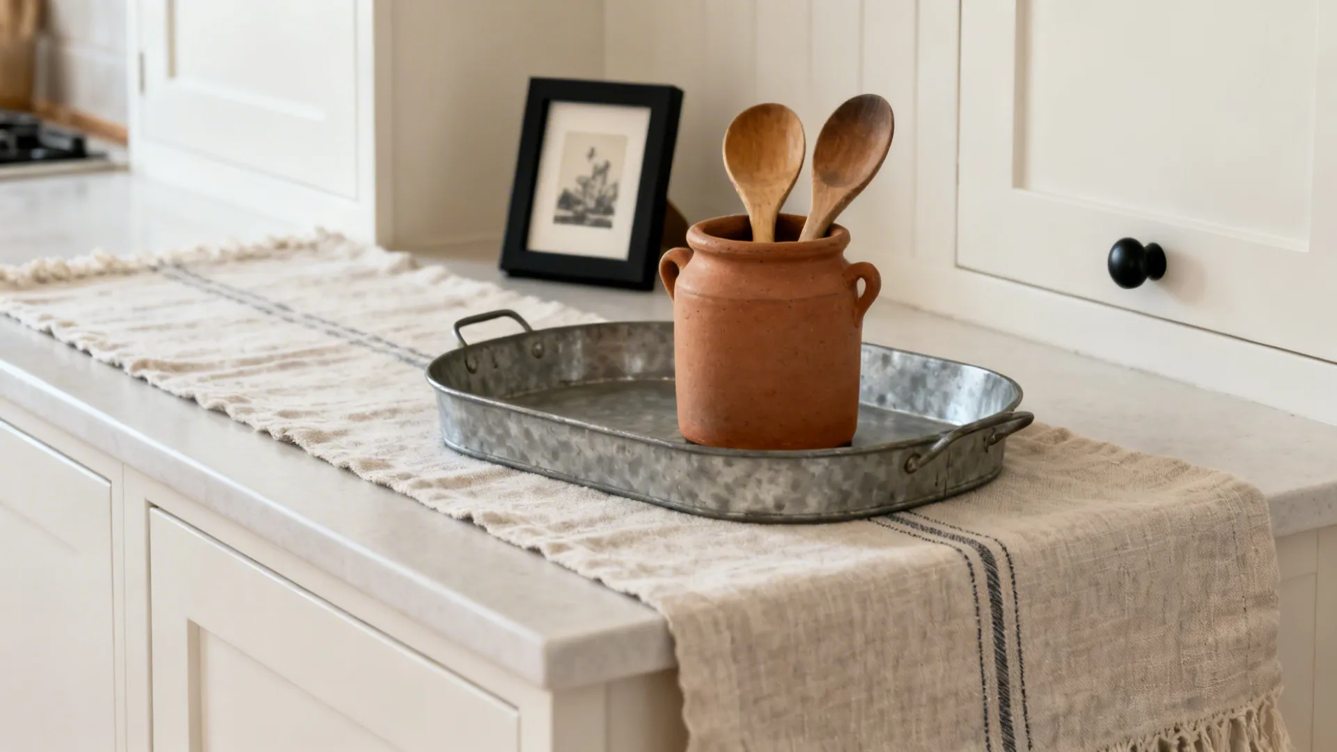 Macro vignette with a linen runner, zinc tray, clay crock and wooden spoons, and a black-framed print.