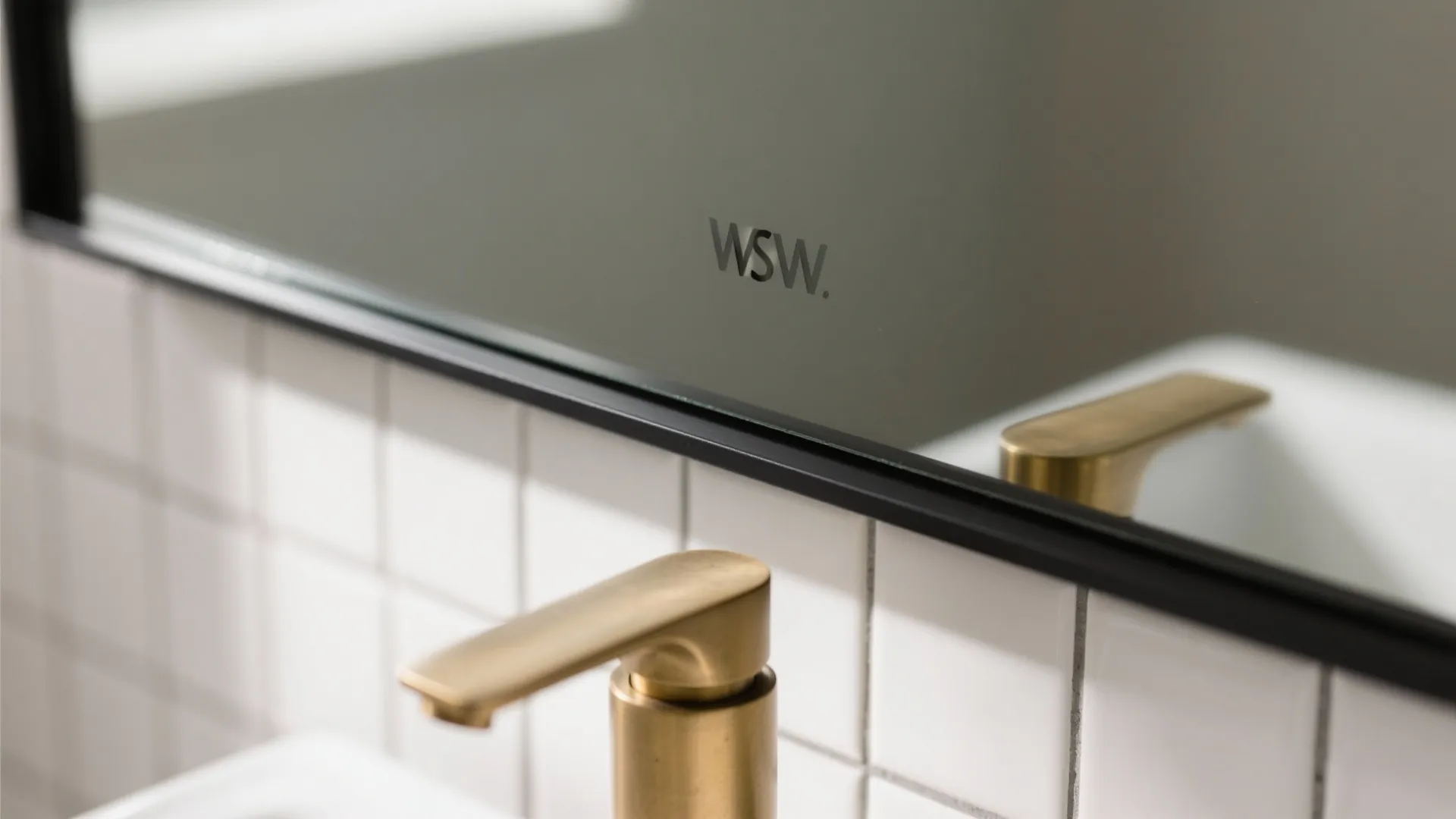 Close-up of bathroom mirror with thin black frame and gold faucet over white tiled wall