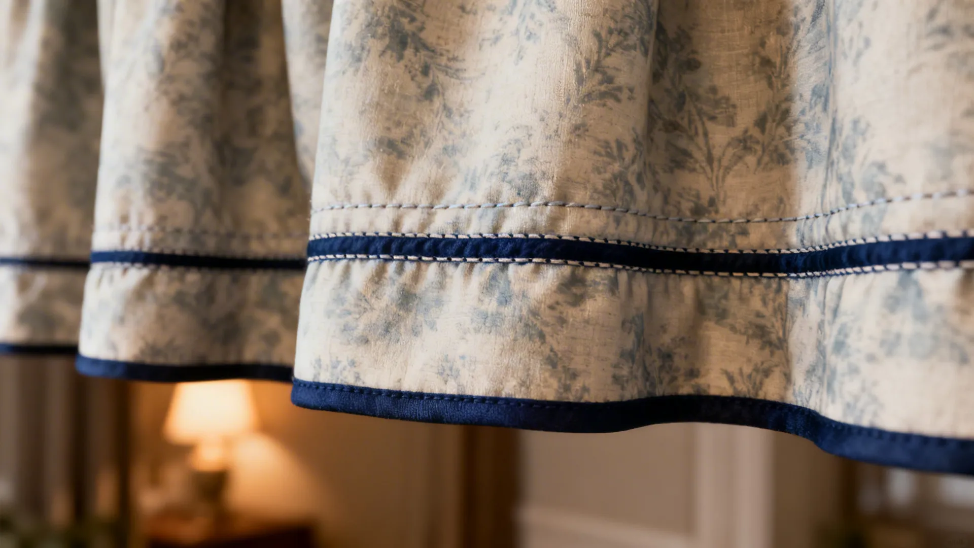Floral pattern curtain with blue trim at the bottom edge near a warm indoor lamp
