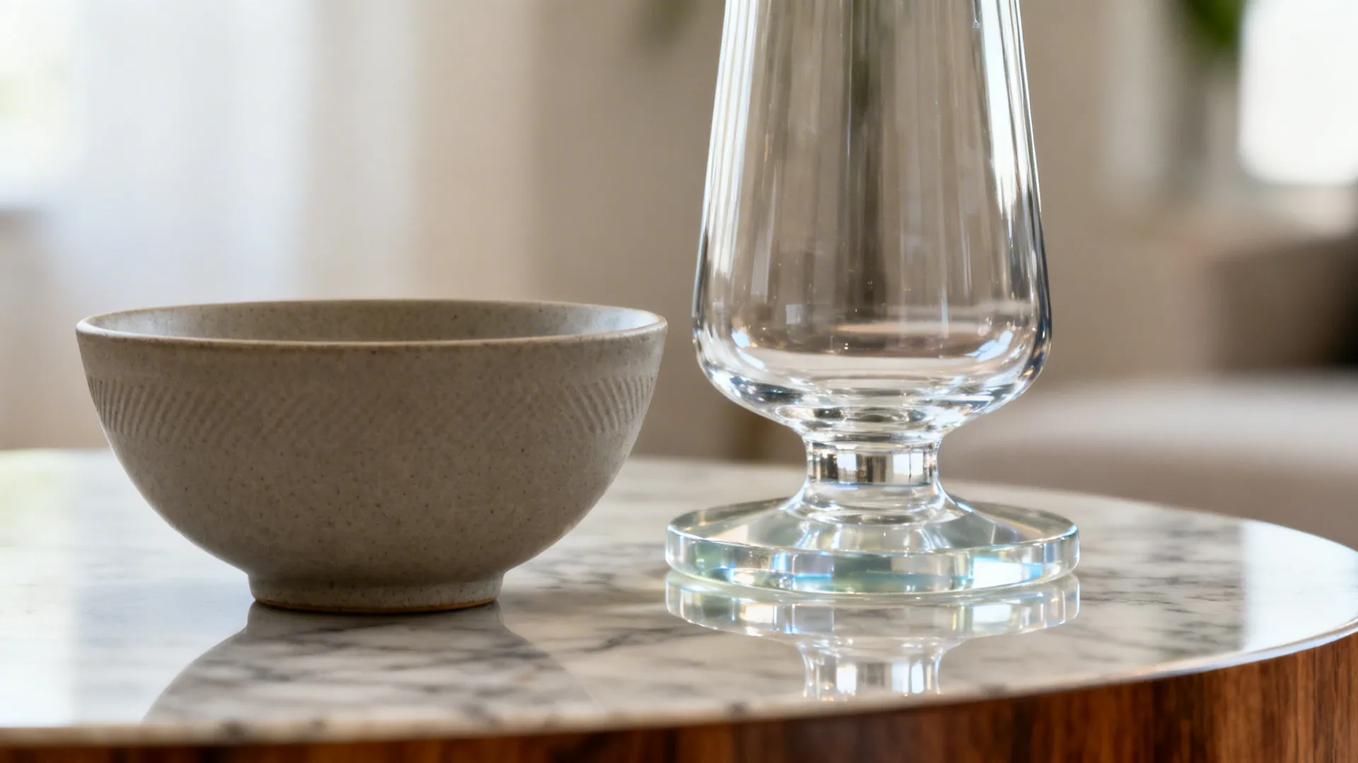 Close-up of a matte ceramic bowl and the base of a tall vase on a centre table with soft daylight reflections.