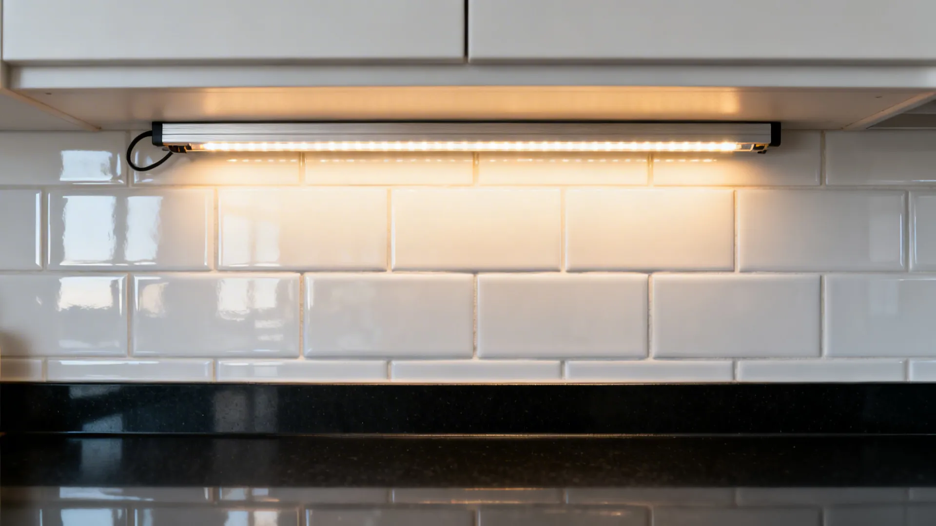 Macro of a warm LED strip in an aluminum channel lighting glossy white tile above a matte black counter.