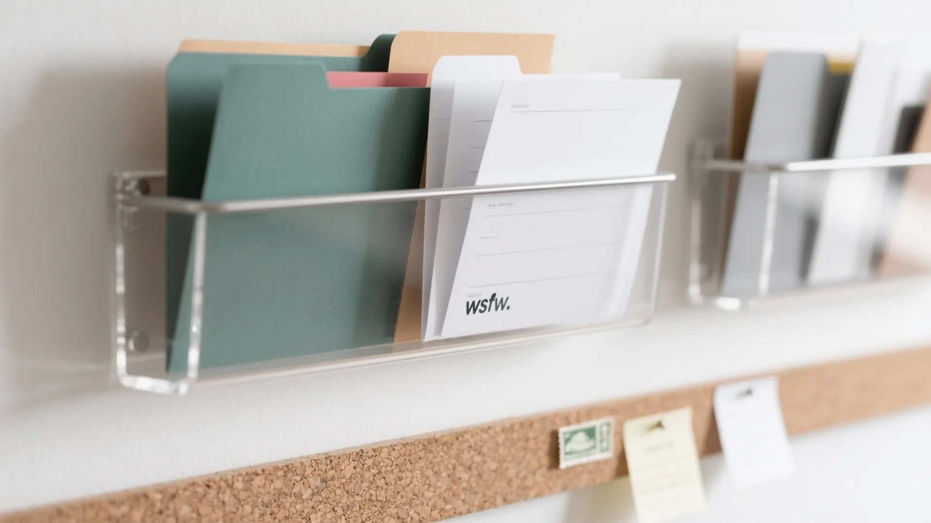 Clear plastic wall mounted organizer holding colorful paper folders and documents above a cork pin board