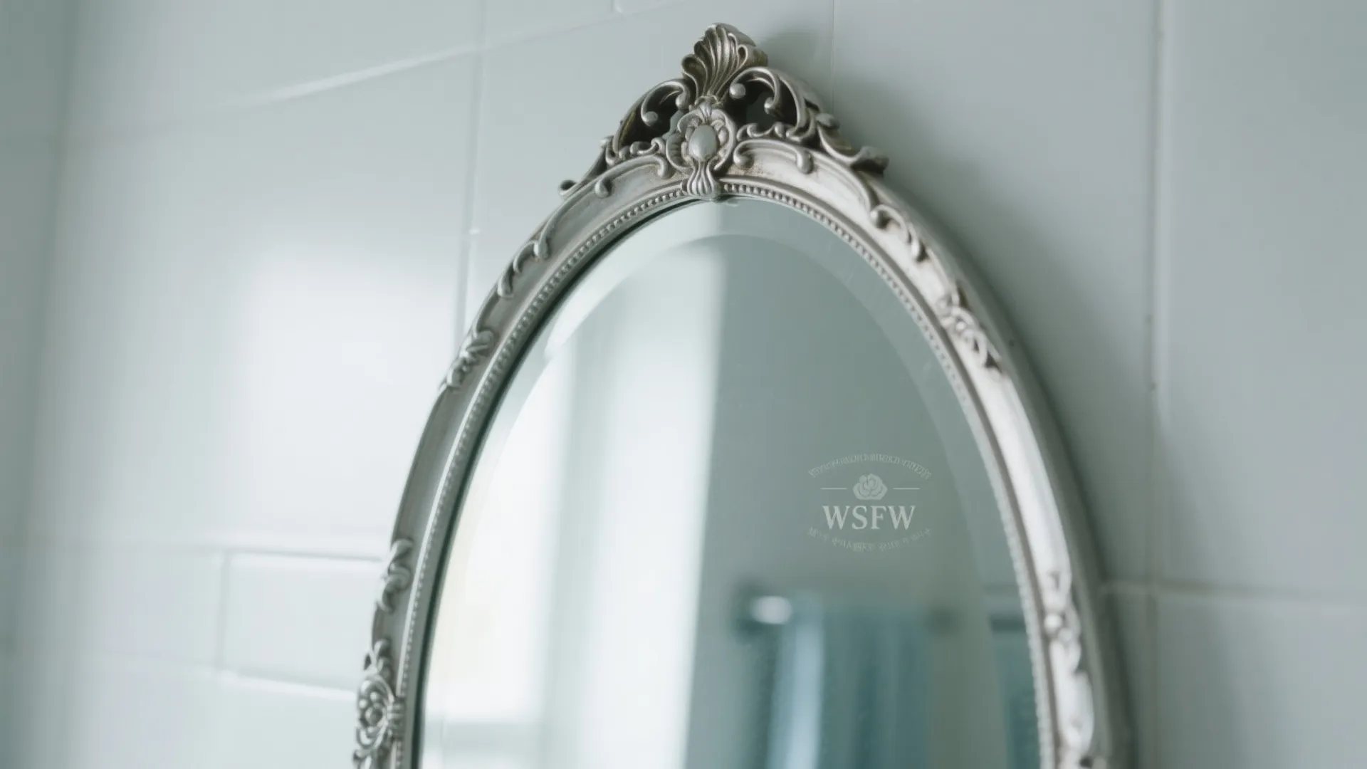 Close up of an oval mirror with ornate silver frame hanging on a white tiled wall