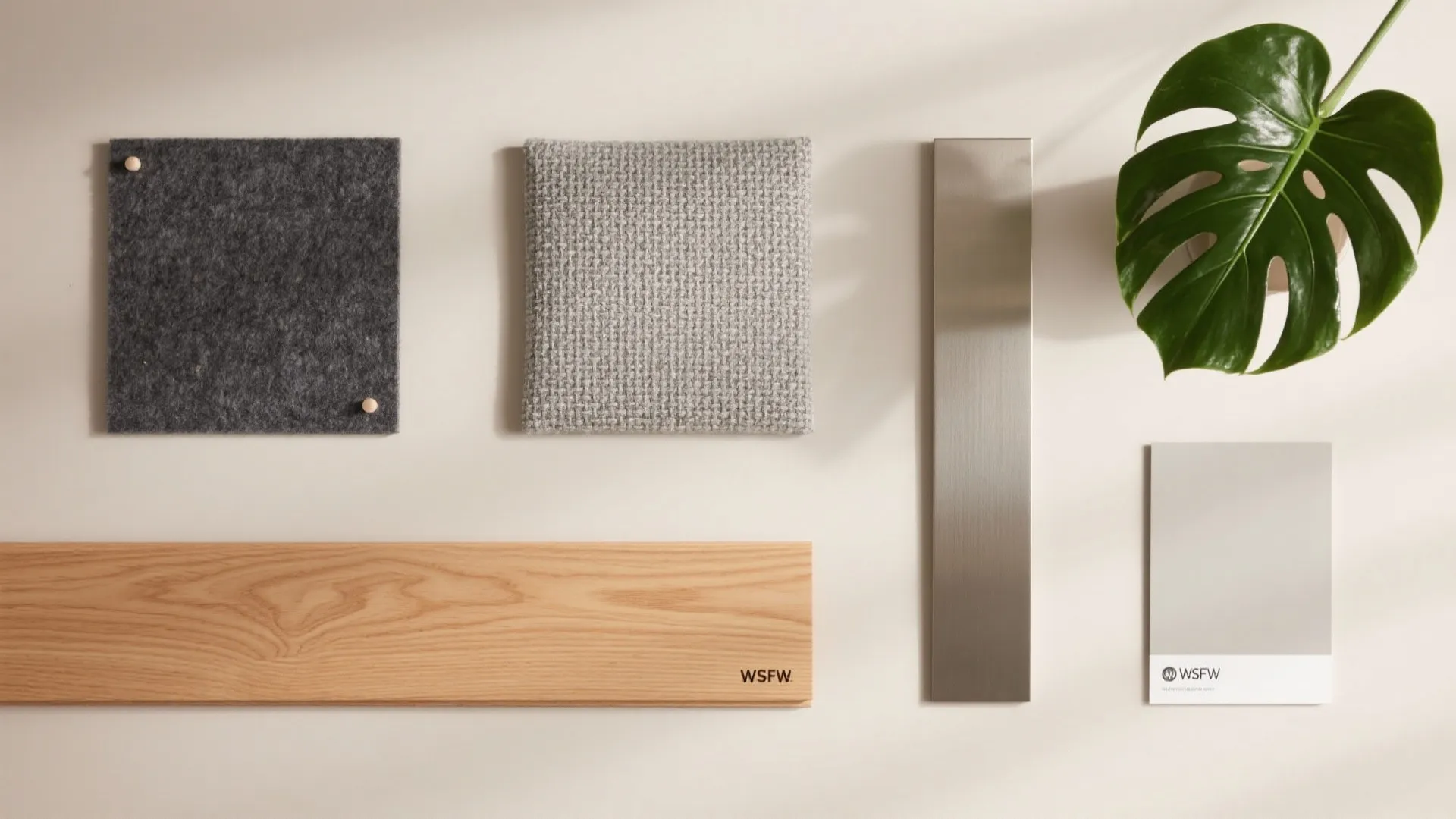 Flat lay of maple laminate, charcoal felt, soft gray upholstery, brushed metal, and a green leaf on a neutral surface.