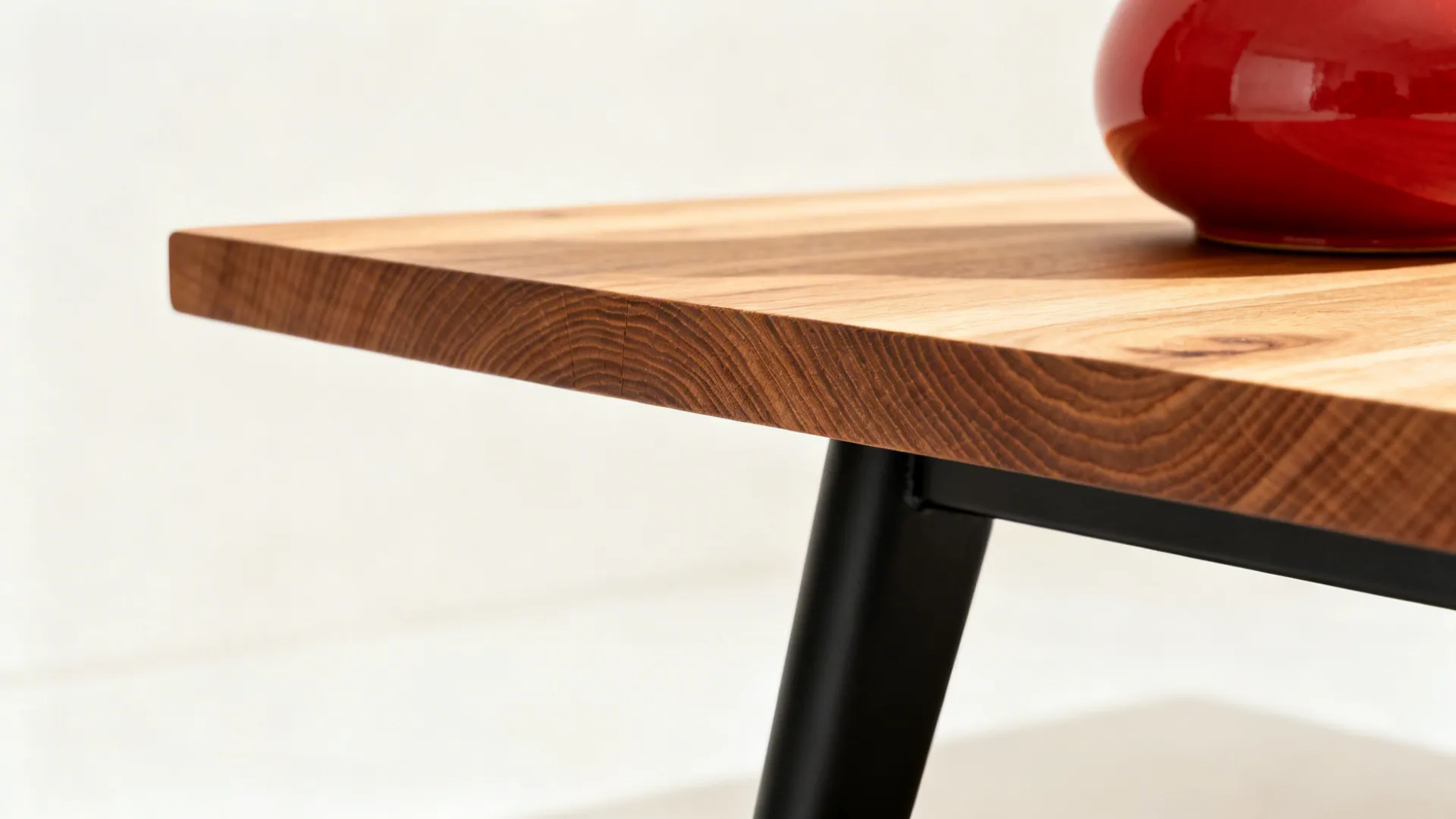 Macro of ash wood table edge, matte black leg, and a red ceramic vase on a warm white backdrop.