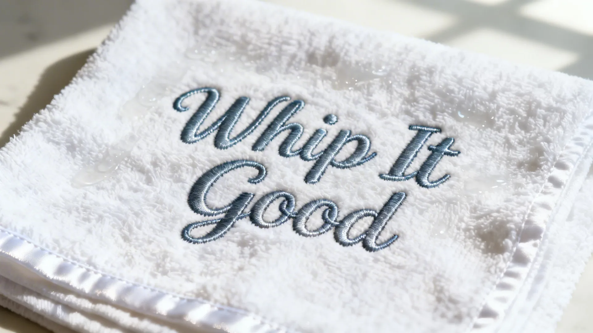 Close up of a white towel featuring blue embroidered cursive text saying whip it good