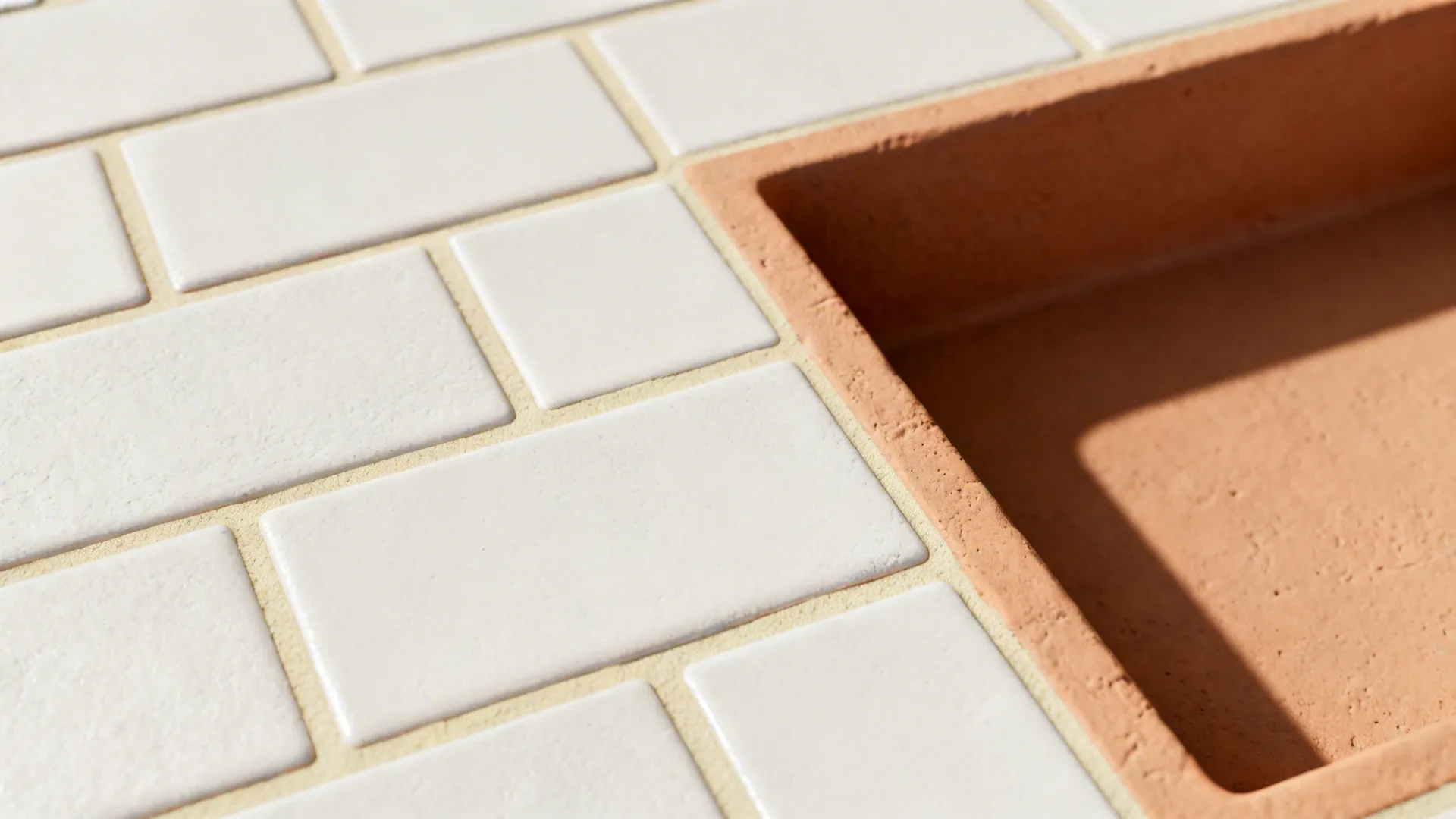 Macro of off-white tile and light-beige epoxy grout with clay-toned niche edge in soft daylight.