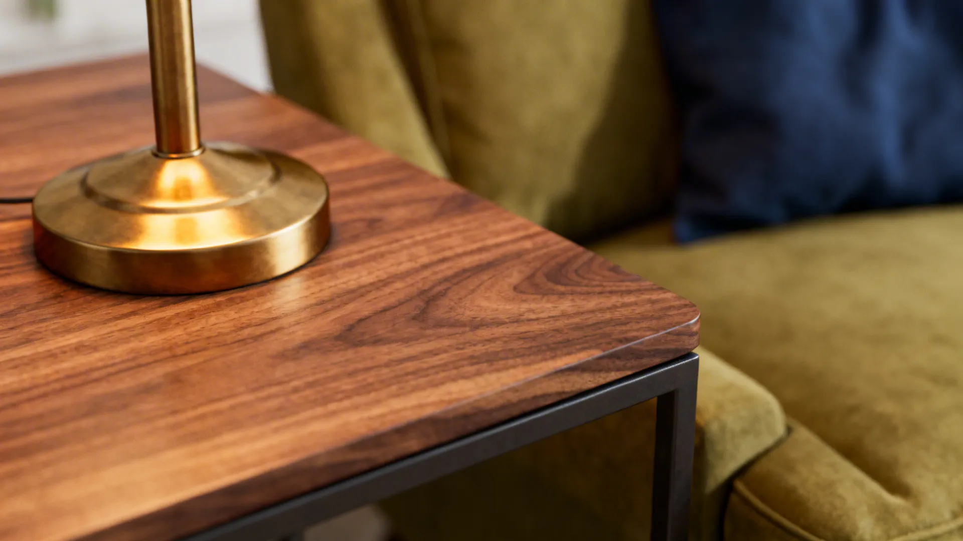 Close-up of walnut table grain and brushed brass lamp near olive upholstery and navy textile.