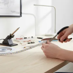 Hands using scissors to cut white LED light strip on clean wood desk with tools